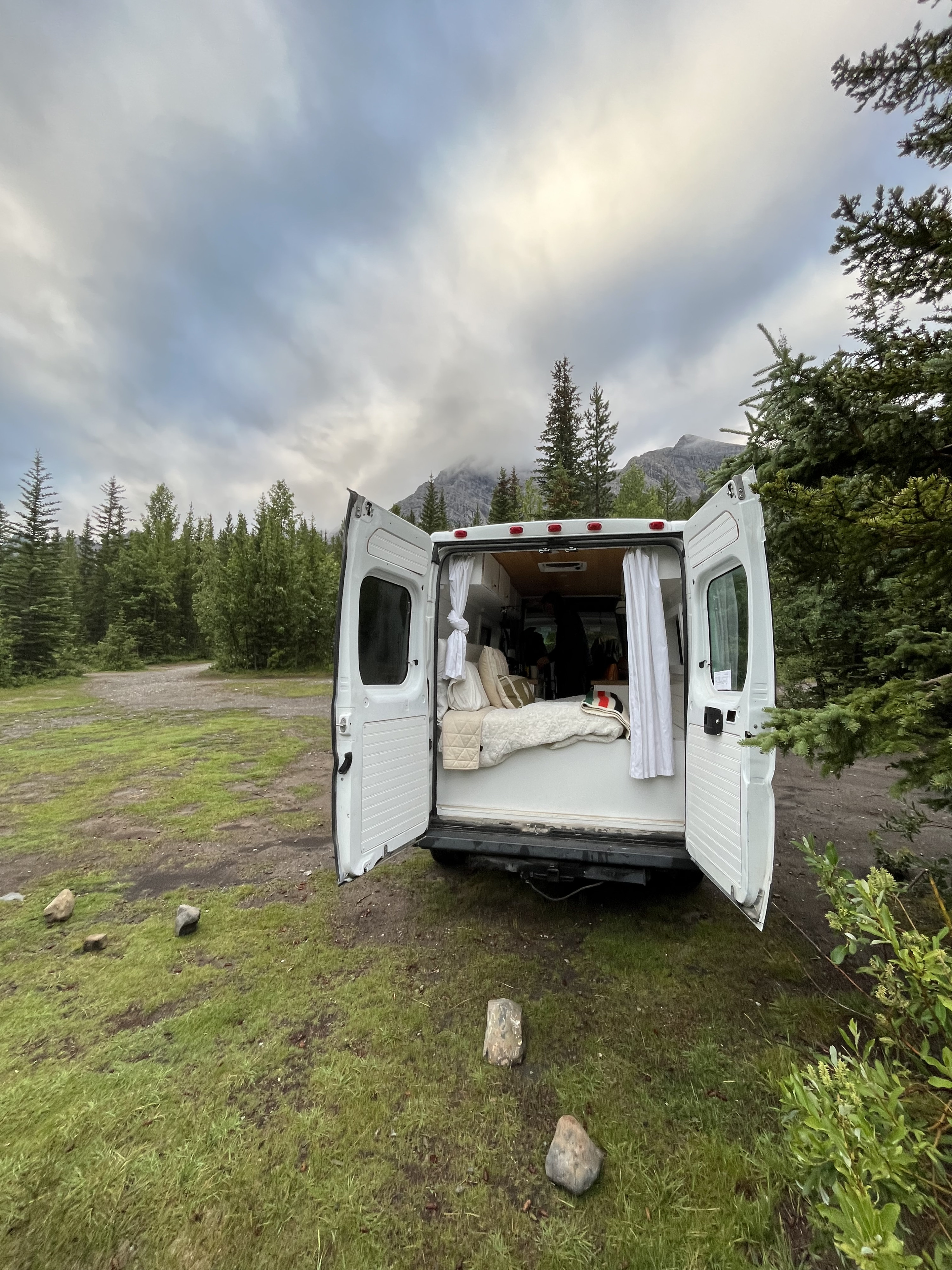 Cozy RV rental with back doors open, revealing a comfy bed. Parked amidst evergreen trees and mountains under a cloudy sky.. Dodge Sprinter 2014