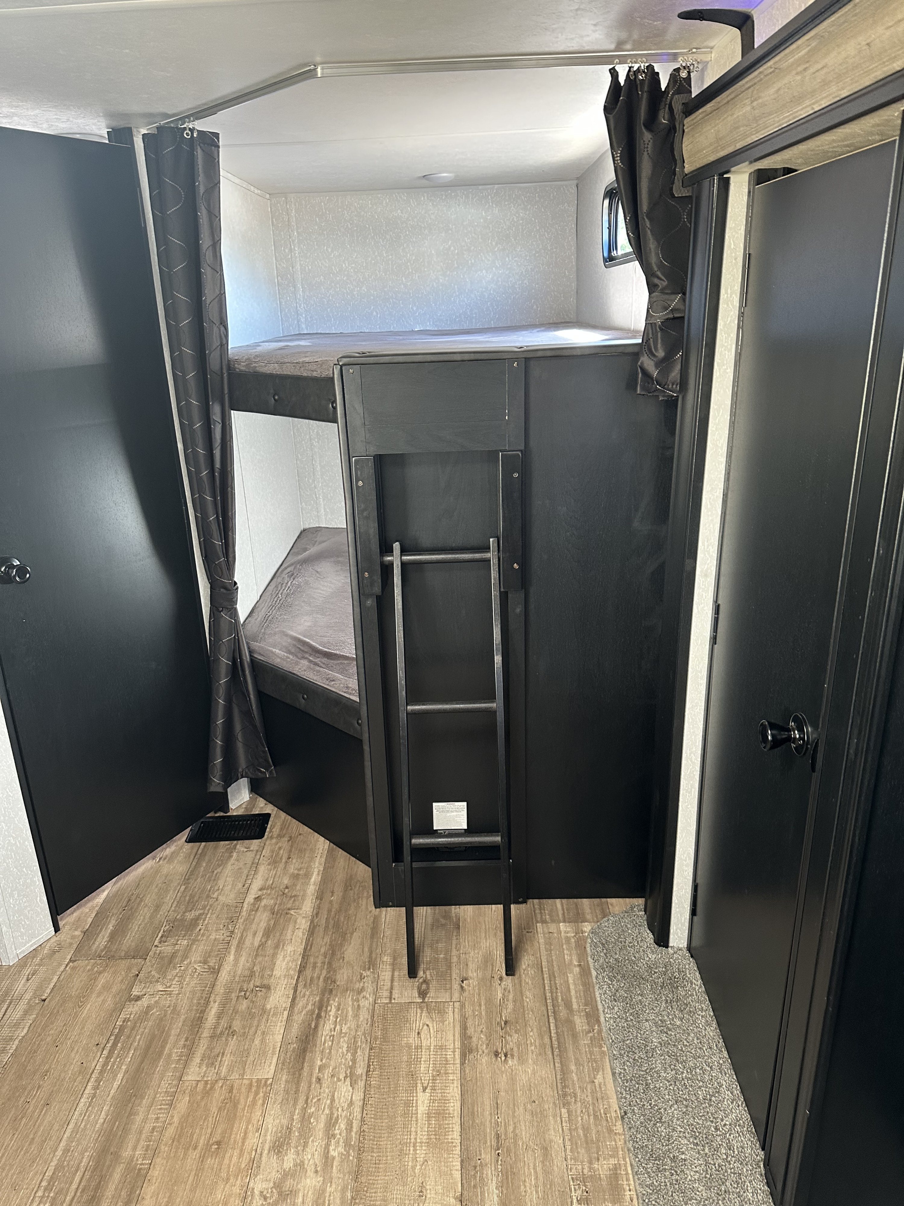 Inside an RV rental: view of the bunk beds with a privacy curtain, ladder, dark doors, and wood-look floor.. Forest River Cherokee Cherokee Black Label 2022