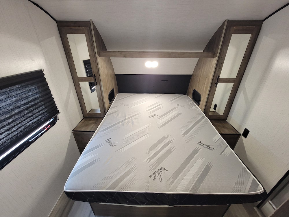 RV rental bedroom featuring a large mattress, overhead cabinets, and a window with blinds, ready for your next adventure.. Heartland RVs Pioneer 2022