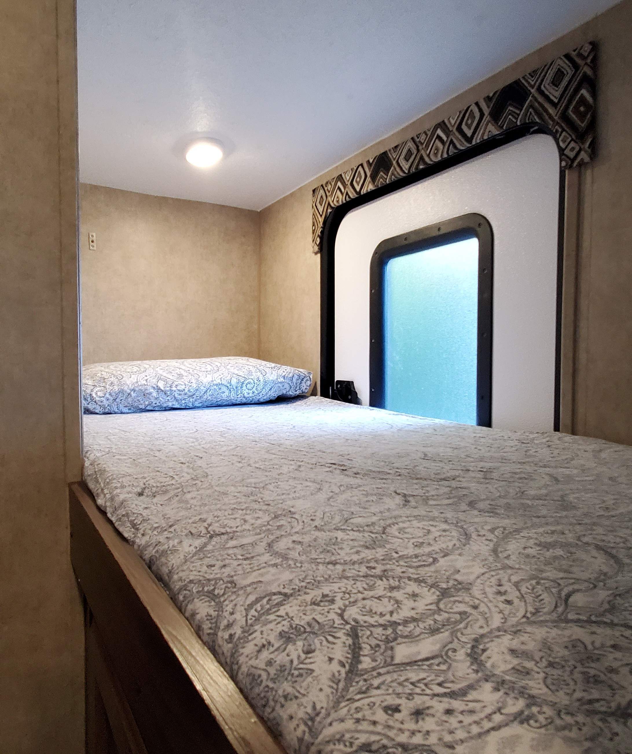 Cozy bunk bed in an RV rental, featuring a patterned duvet, pillow, window, and warm ceiling light, all set against beige walls.. Coachmen Other 2018
