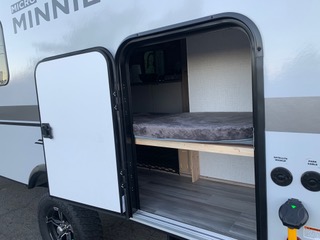 This RV rental shows its open door revealing a compact bunk bed inside. Exterior utility hookups are visible. Ideal for travel & camping adventures.. Winnebago Minnie Winnie 2021
