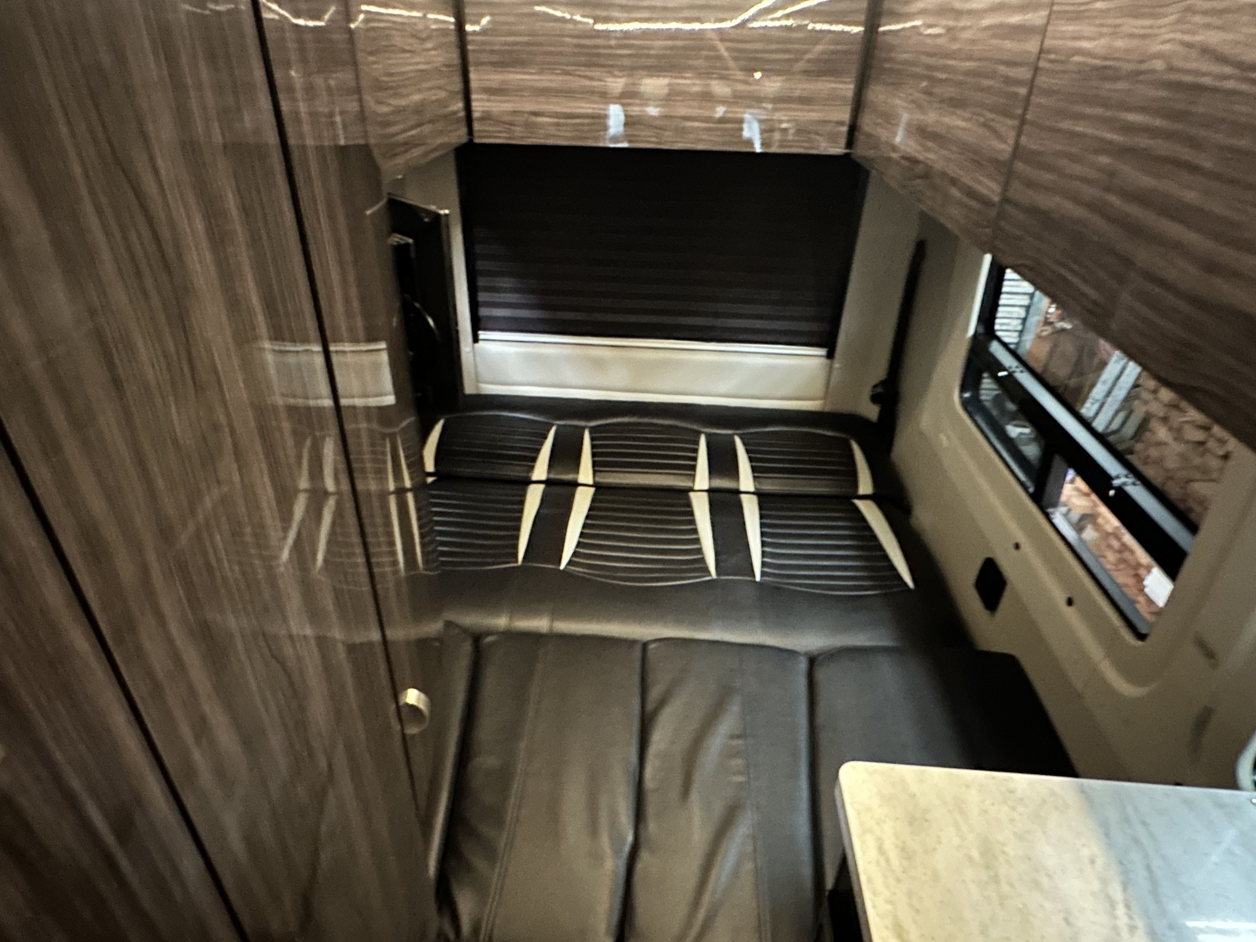 Interior of an RV rental: black, modular seating converts to a bed, with wood paneling and window view. Compact travel comfort.. Winnebago Era 2017
