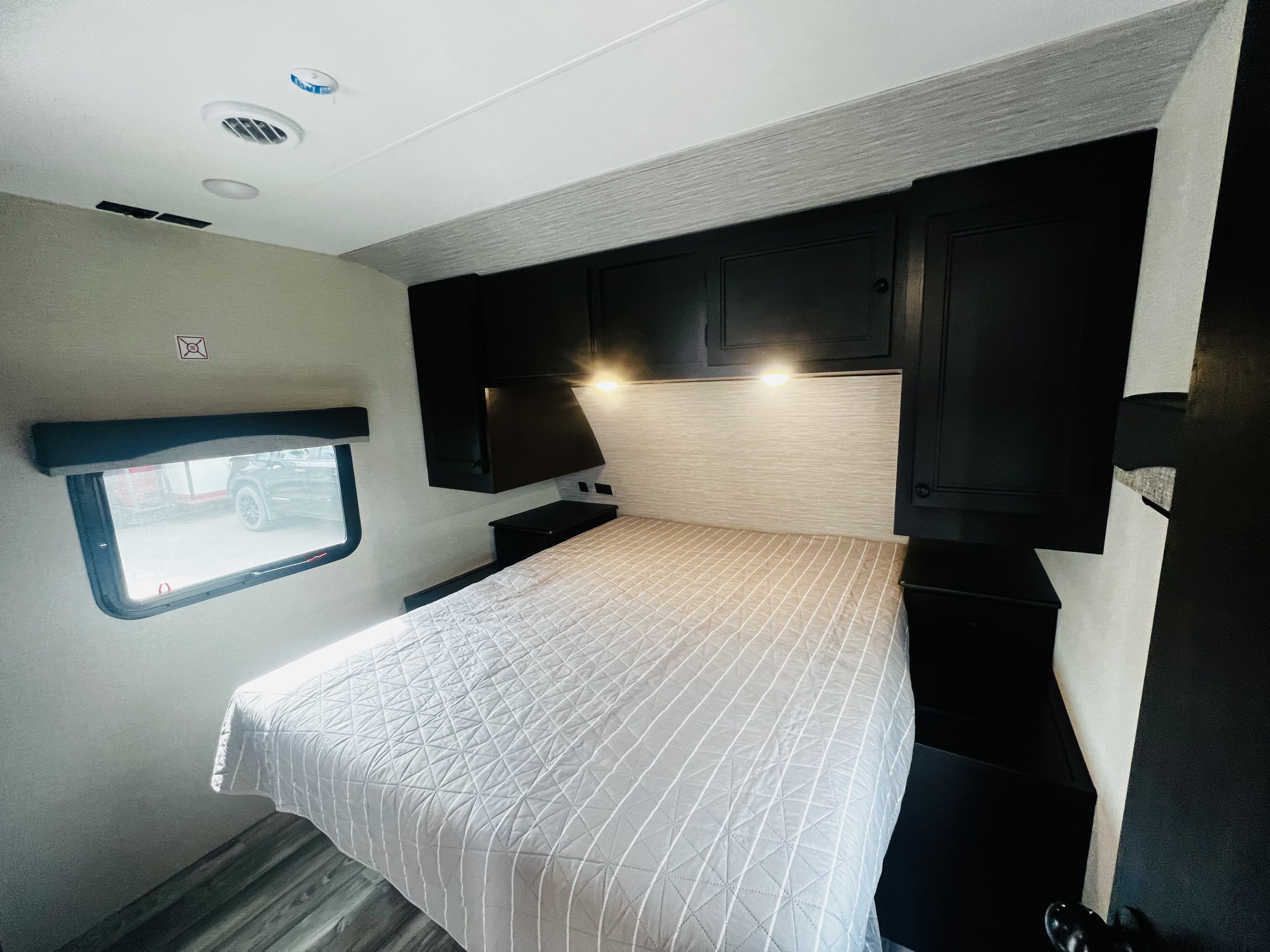 Bright RV rental bedroom: cozy bed with quilted spread, black overhead cabinets, reading lights, and side window.. Highland Ridge RV Range Lite 2022