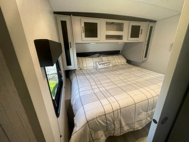 RV rental's compact bedroom: cozy bed with plaid comforter, overhead cabinets, and side window.. Heartland RVs North Trail 2022