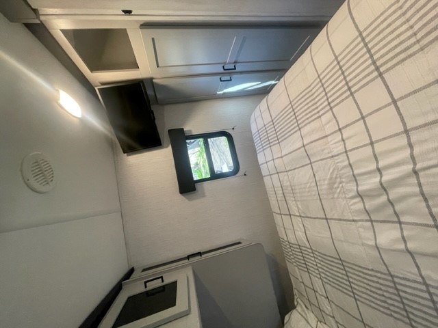 Inside a compact RV rental bedroom: bed with plaid cover, mounted TV, window, and overhead storage.. Heartland RVs North Trail 2022