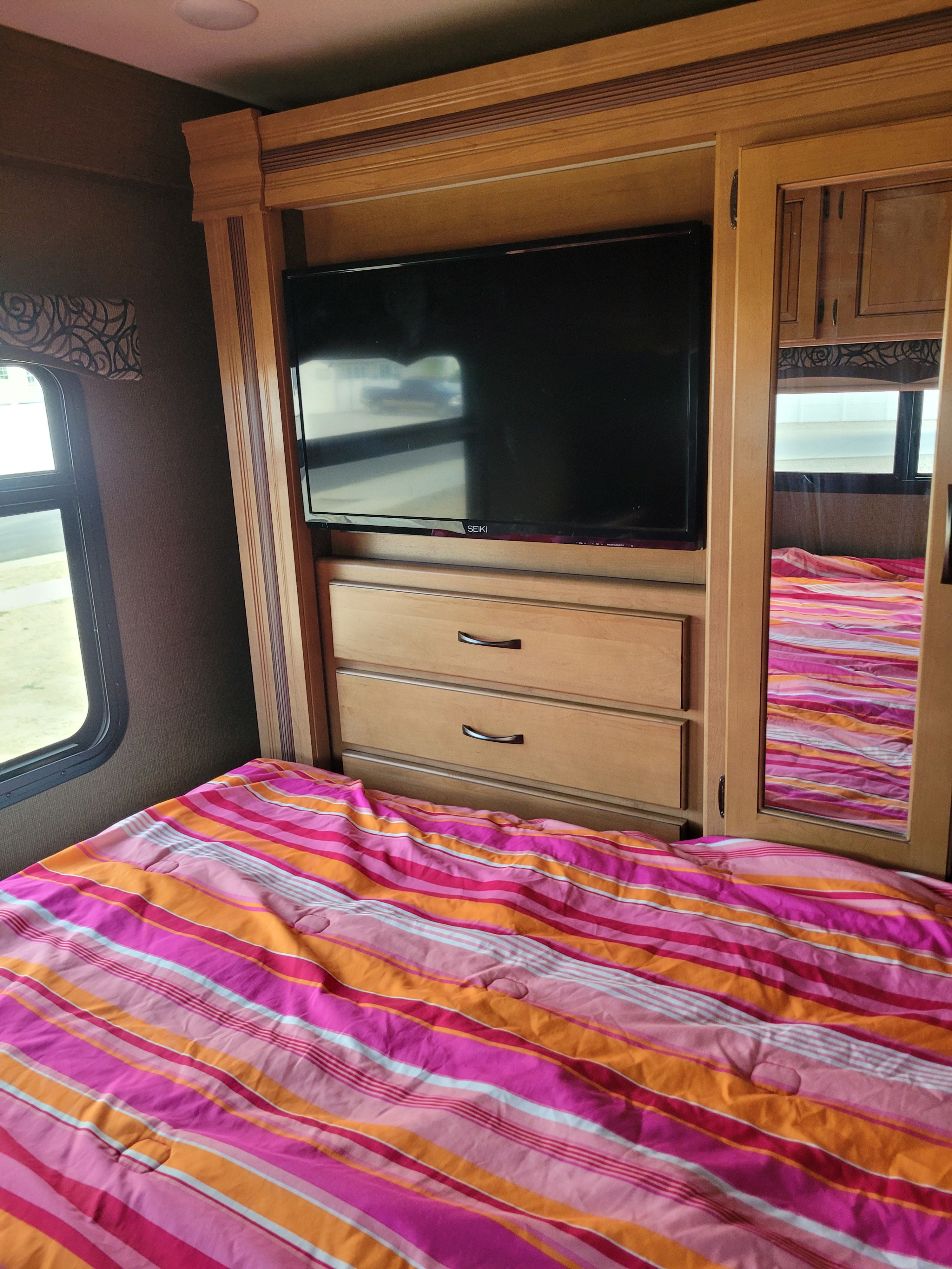 RV rental bedroom: Vibrant striped bedding on bed next to built-in TV, dresser, and mirrored wardrobe, offering cozy and functional space.. Thor Four Winds 2017