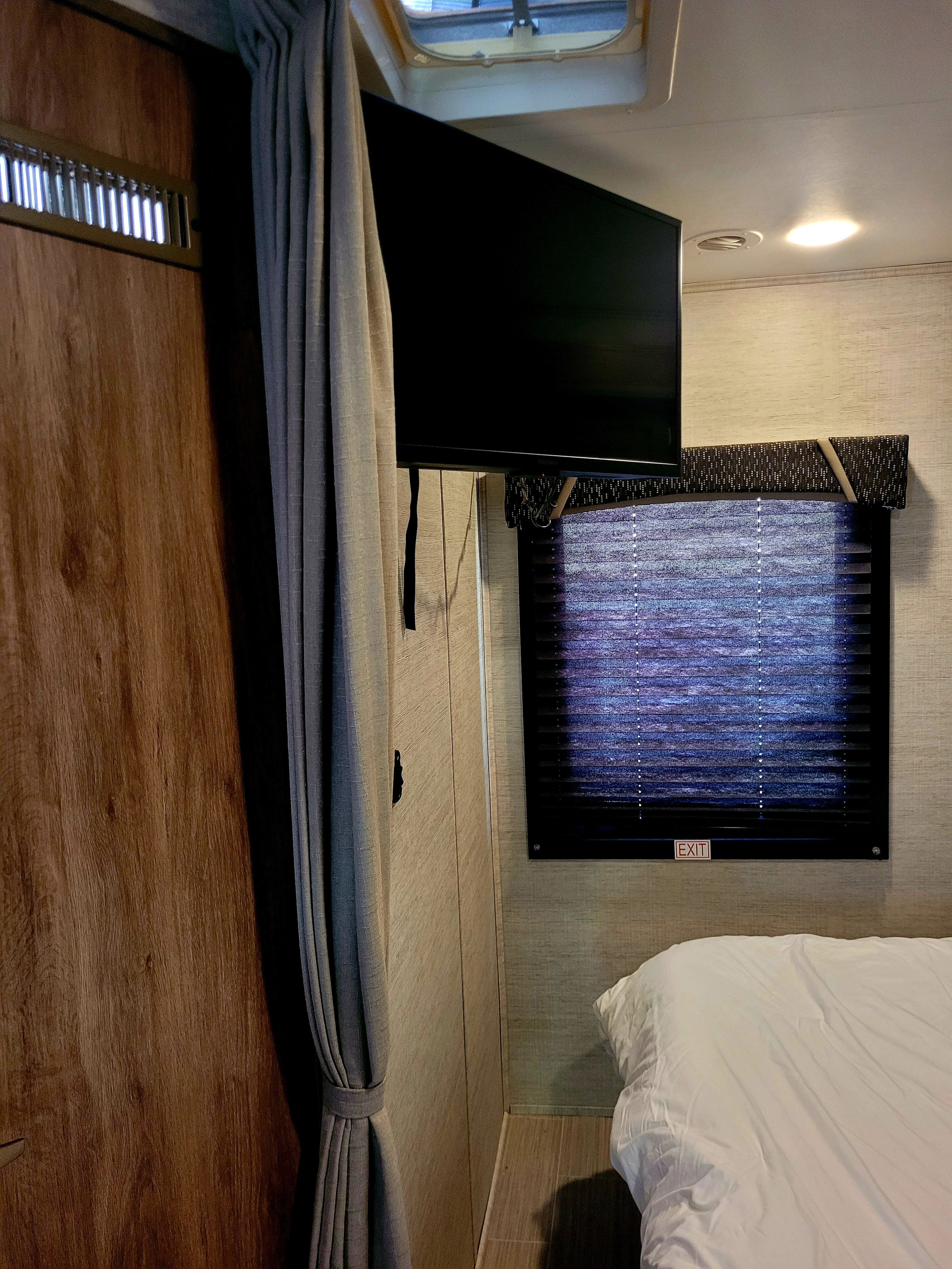 Inside an RV rental bedroom: a wall-mounted TV, window with an 'EXIT' blind, and a white-sheeted bed.. Jayco Redhawk SE 2021