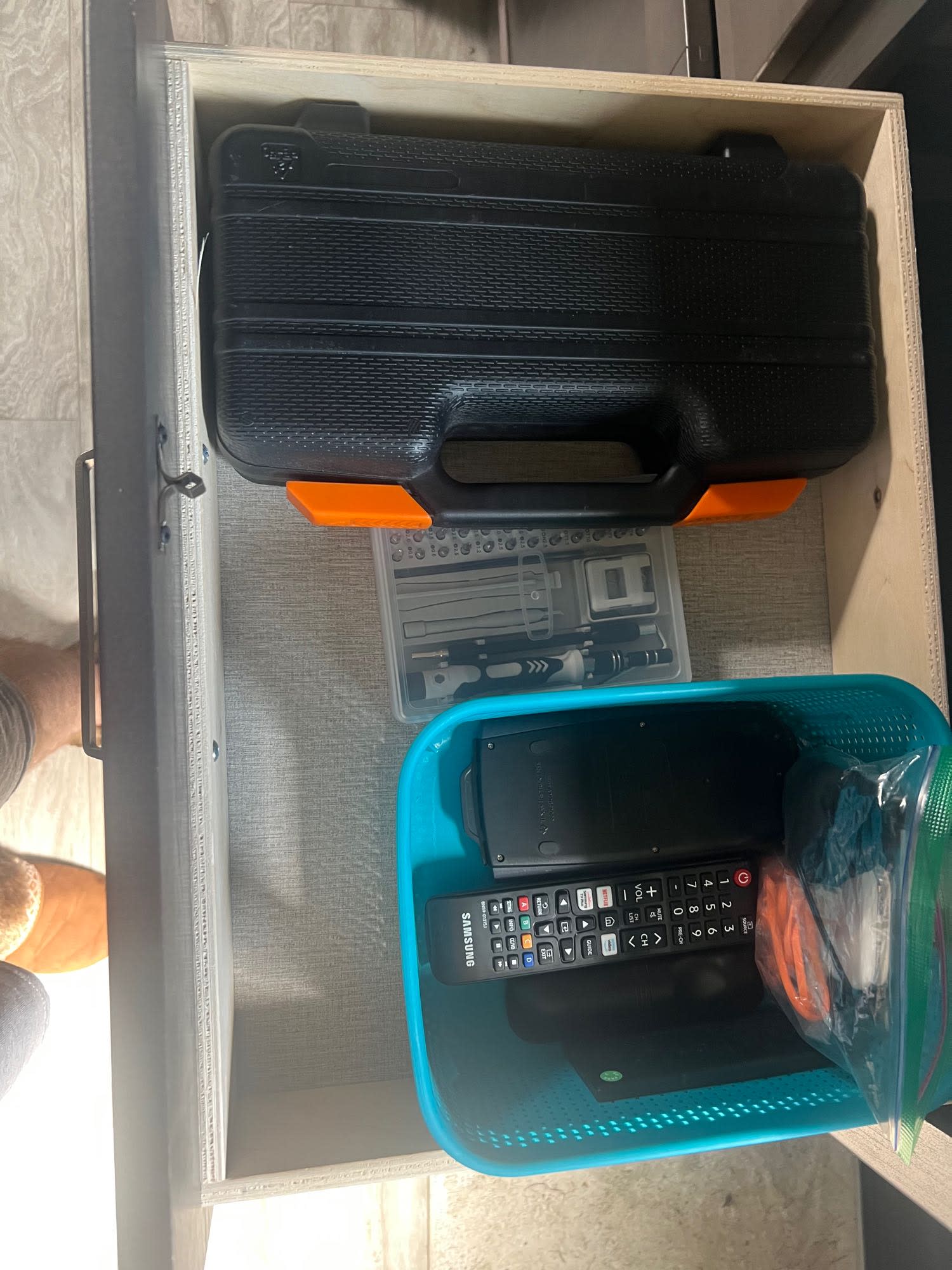 A pulled-out drawer in an RV rental reveals a tool case, precision kit, and a basket with electronics like remotes.. Grand Design Transcend Xplor 2022