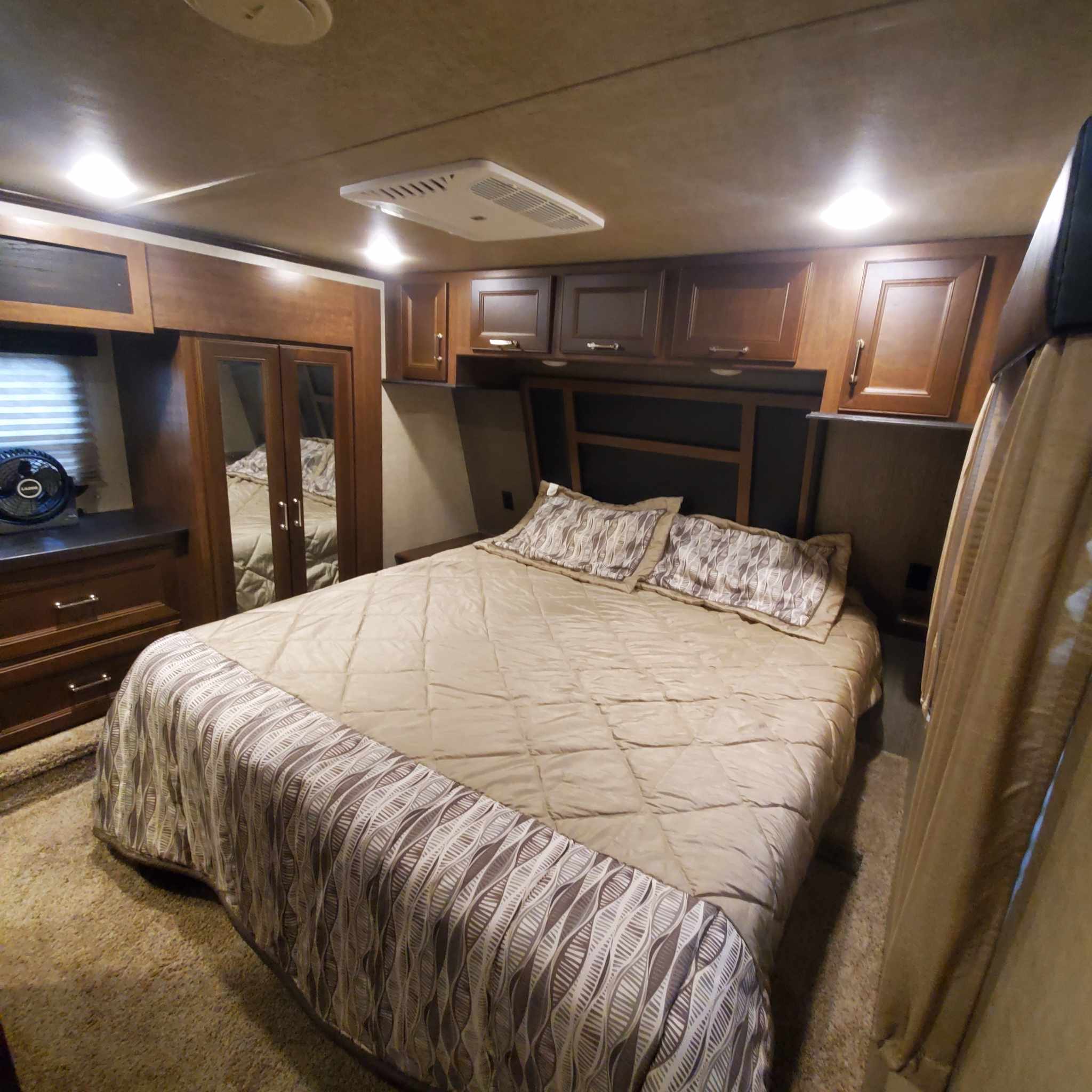 This RV rental bedroom features a cozy bed with patterned bedding, mirrored closet, overhead cabinets, and soft lighting.. Keystone RV Fuzion Toy Hauler 2016