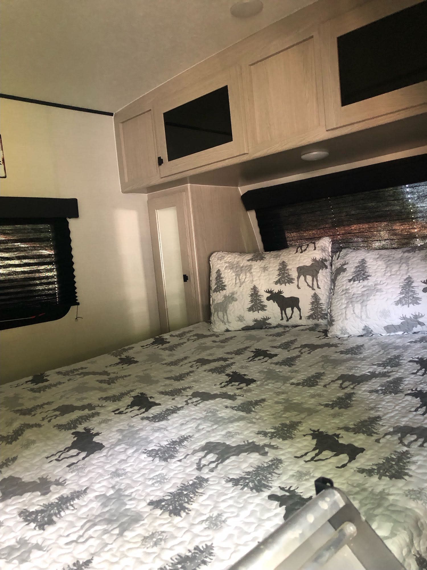 Cozy RV rental bedroom with moose-themed quilt, pillows, overhead cabinets, and window blind. Ideal for nature lovers.. Coachmen Apex Nano 2023