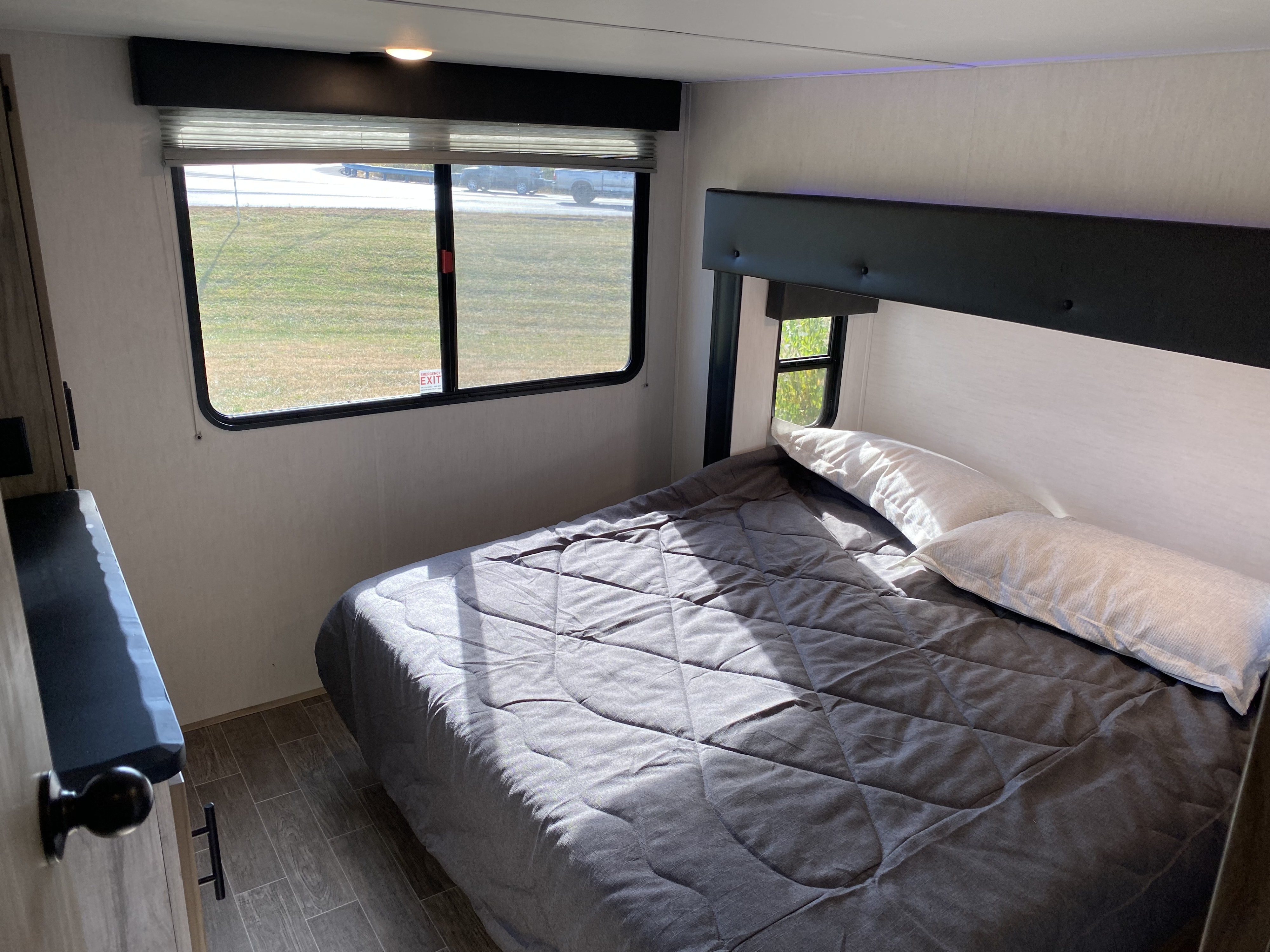 Bright RV rental bedroom: gray bed, pillows, sunlit window view of grass & road. Comfortable travel living.. Forest River Cherokee Arctic Wolf 2022