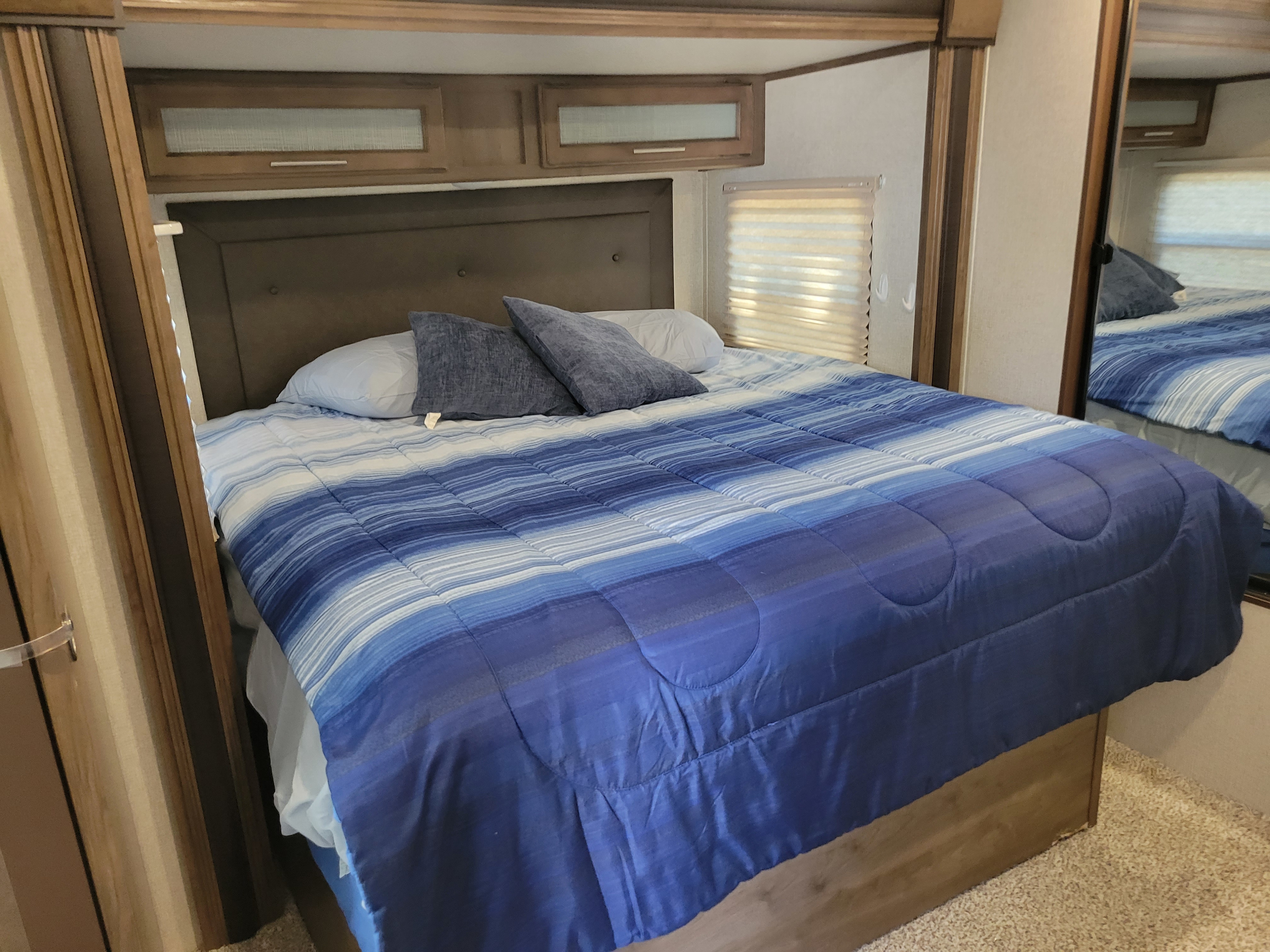 Inside an RV rental, a comfy bedroom features a blue striped comforter, dark headboard, overhead cabinets, and a window with blinds.. Keystone RV Cougar 2019