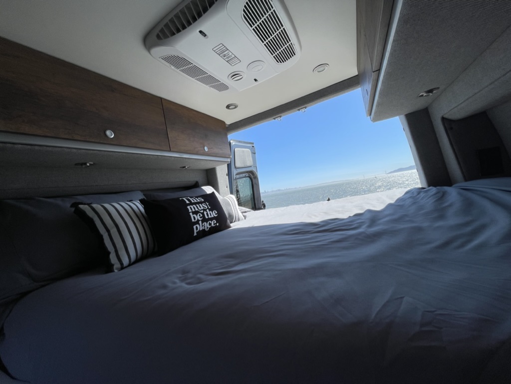 Cozy RV rental interior with a comfy bed offering breathtaking bay views from the open rear doors, perfect for relaxing travel.. Storyteller Overland Storyteller Overland Class B 2022