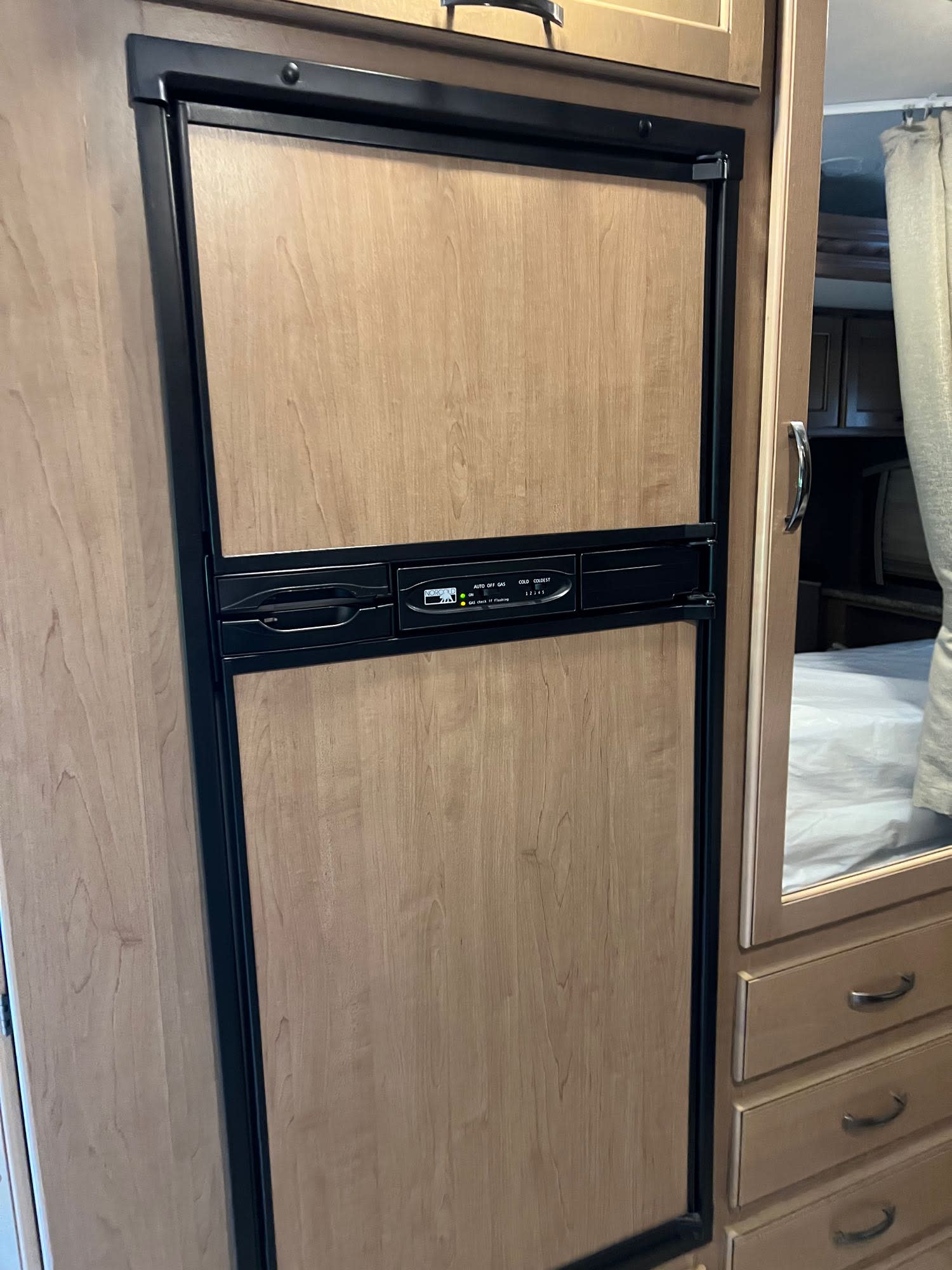 Inside this RV rental, a wooden refrigerator with a control panel and black trim stands next to a mirrored dresser and bed.. Thor Chateau 2018