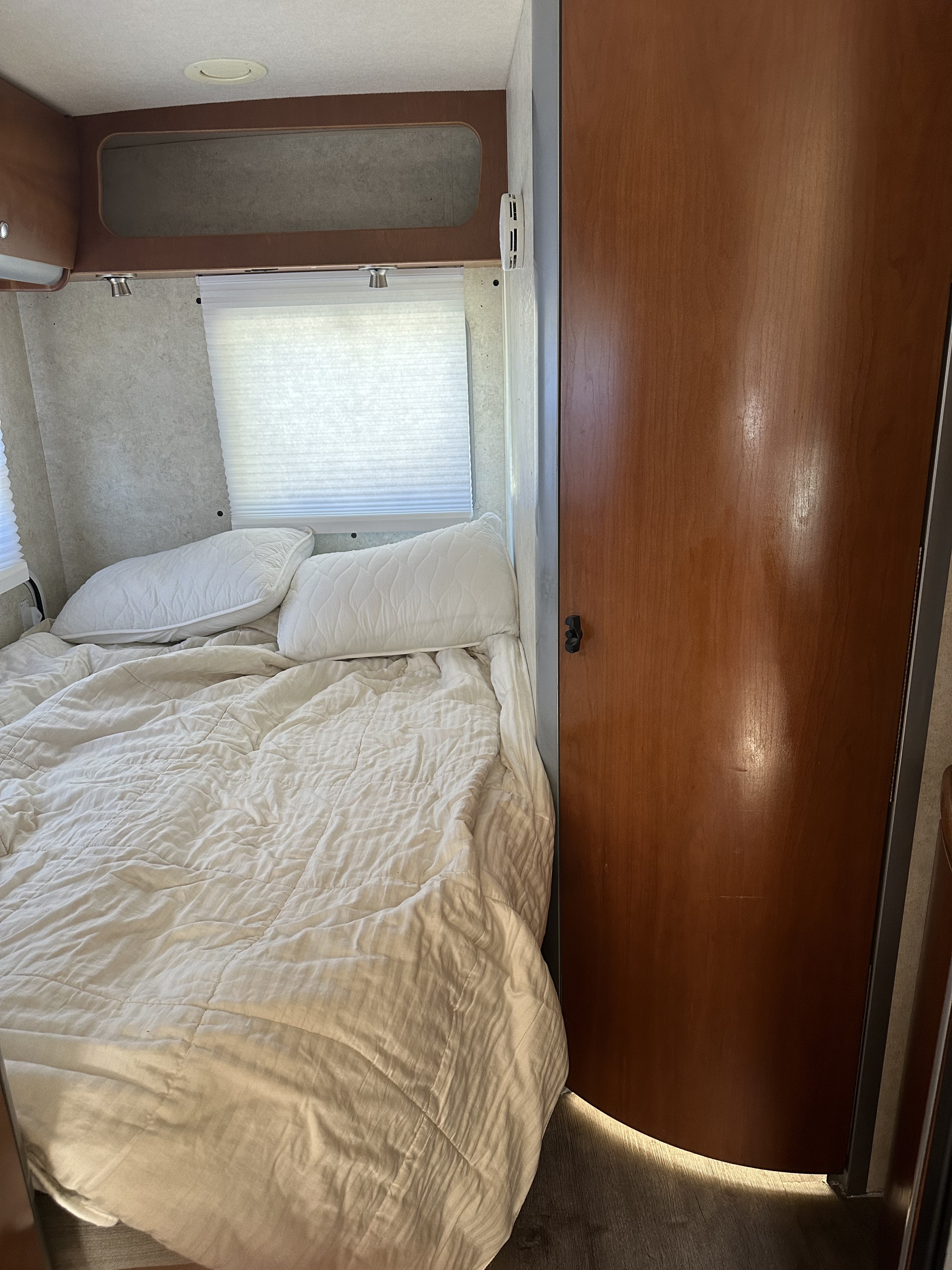 Relax in this clean RV rental bedroom, featuring a comfortable bed with white bedding, windows, and wood accents.. Winnebago View 2006