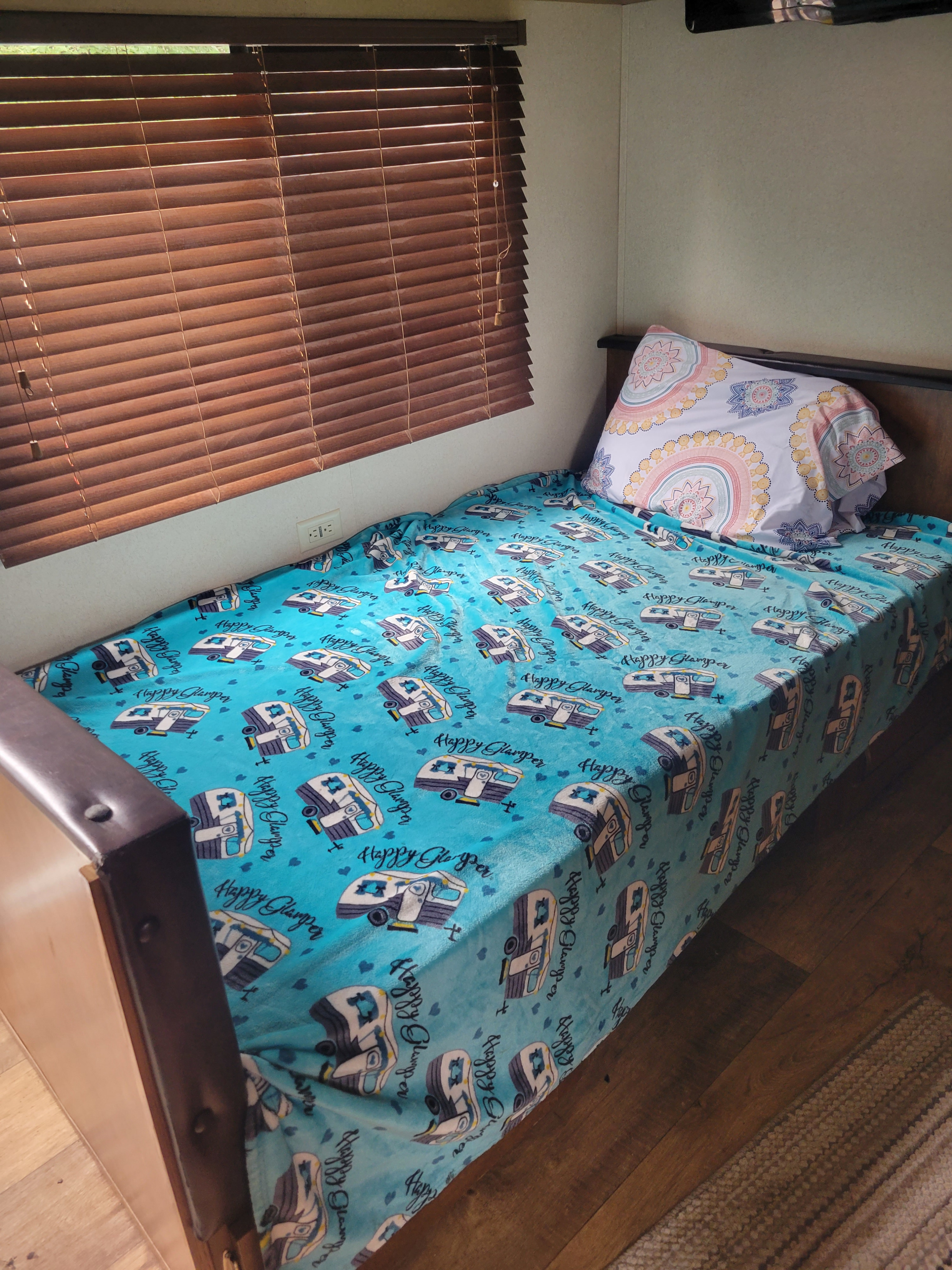 Cozy RV rental bed with a fun camper-themed blanket and decorative pillow, next to wooden blinds.. Forest River Wildwood X-Lite 2017