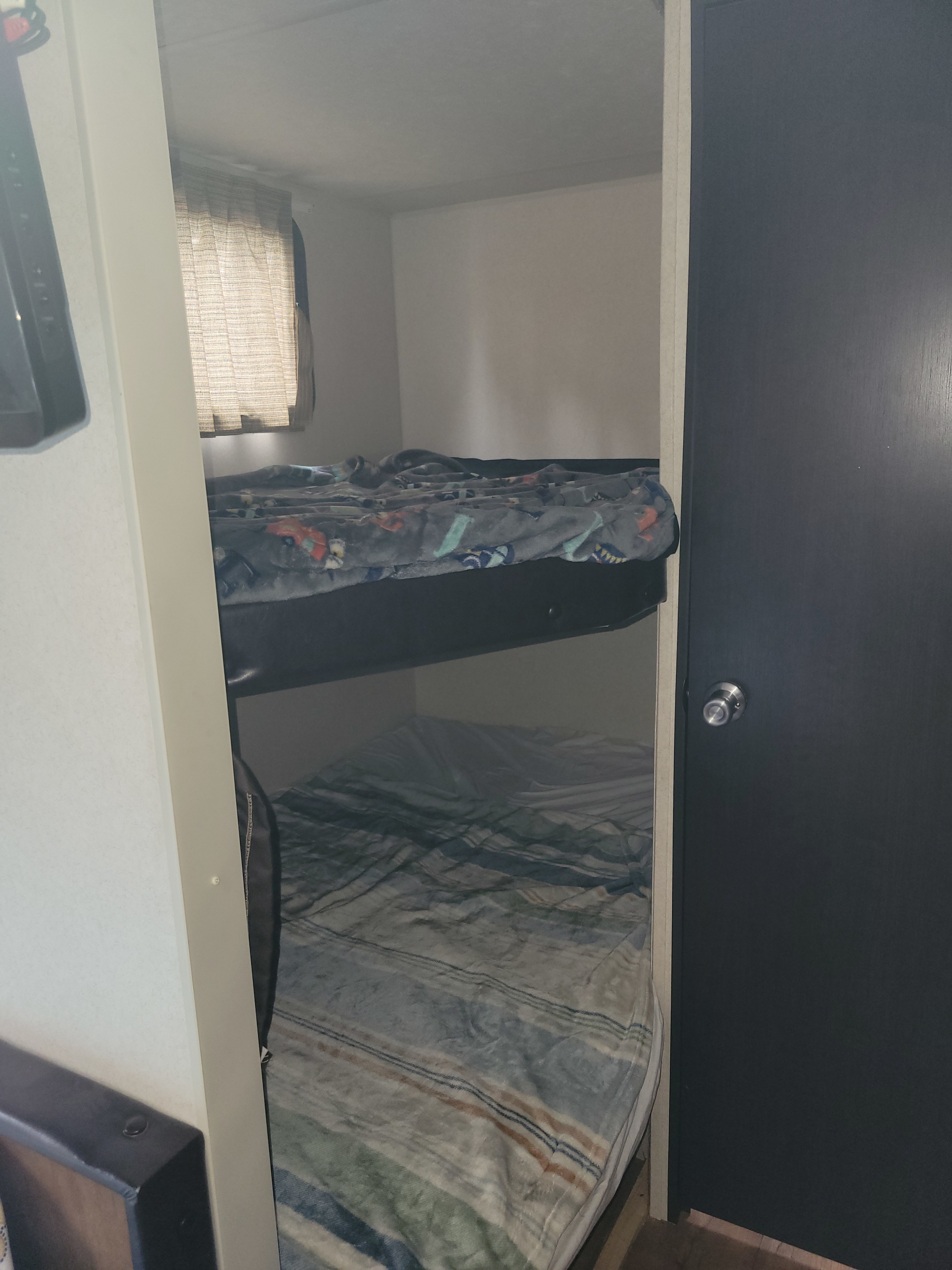 Inside an **rv rental**, showing a small bedroom area with two bunk beds, a window on the left, and a dark door on the right.. Forest River Wildwood X-Lite 2017