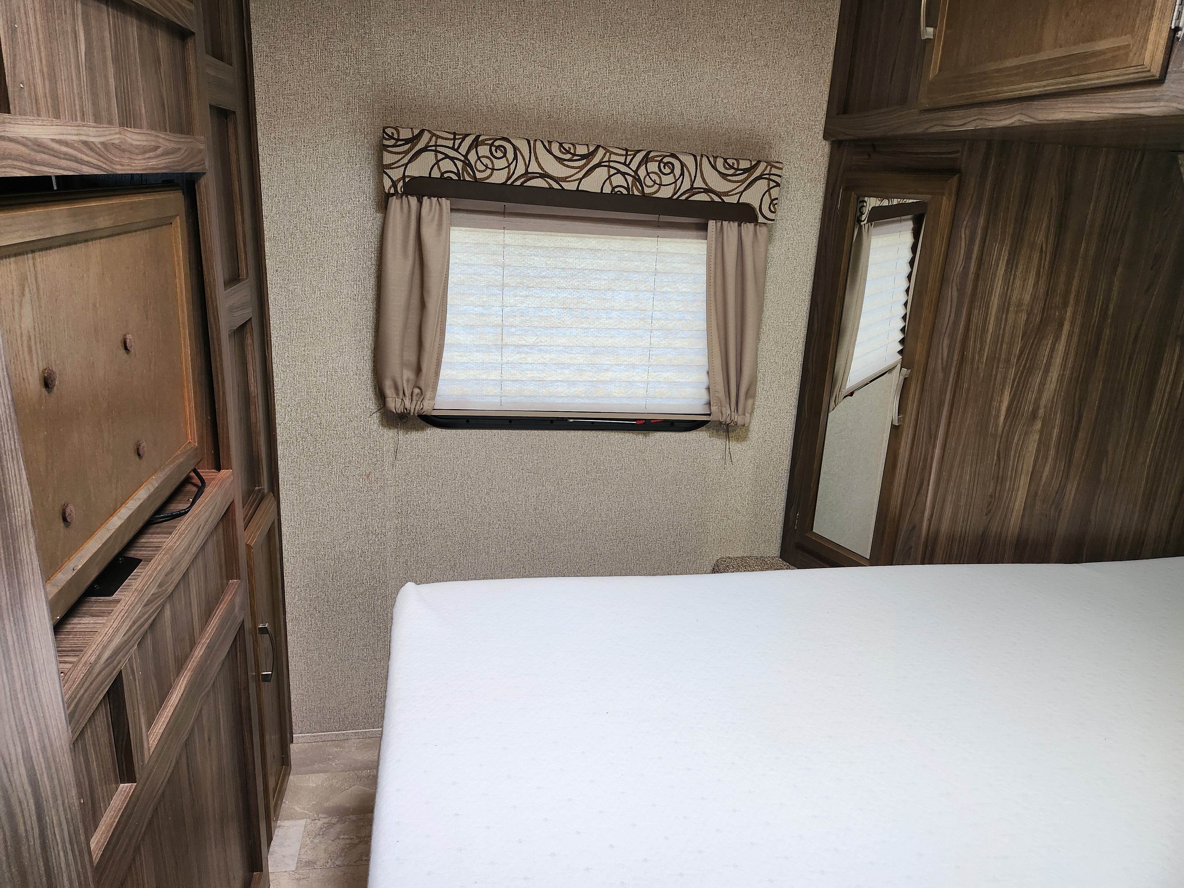 Compact RV rental bedroom with a white bed, window, and rich wood cabinetry, offering functional comfort.. Coachmen Apex 2017