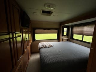 RV rental bedroom: A full-size bed with grey cover, large windows, and built-in wooden cabinets/TV offer a cozy space for your next trip.. Forest River Sunseeker 2011