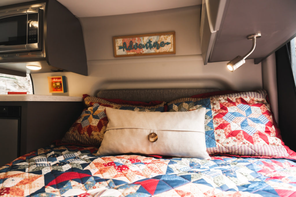 Relax in this cozy RV rental bed! Features a colorful quilt, microwave, and an "Adventure" sign, ideal for road trips.. Thor Sanctuary 2023