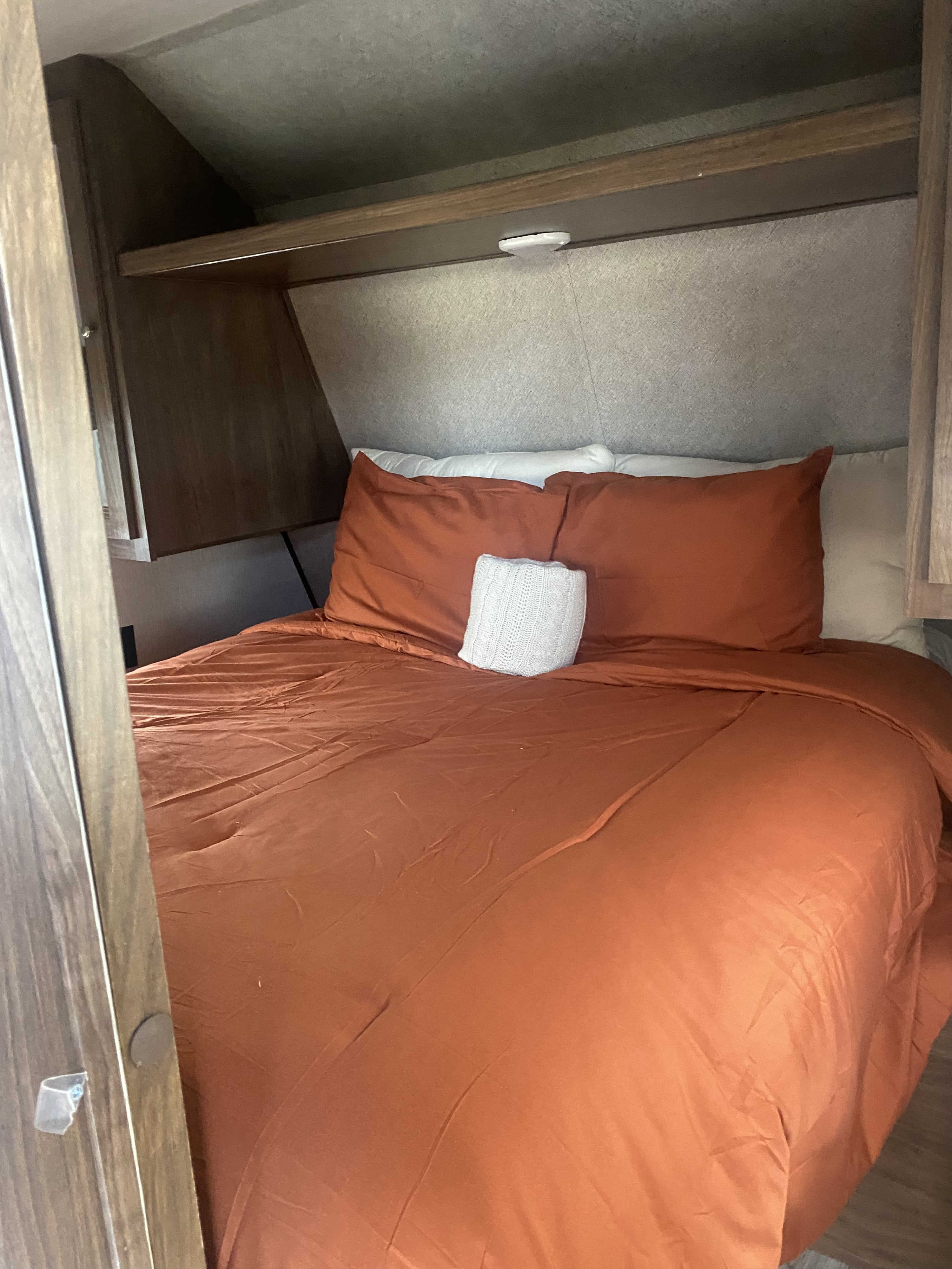 An RV rental bedroom with a neatly made bed, featuring orange bedding and a white pillow, alongside overhead storage and lighting.. Heartland RVs Prowler Lynx 2018