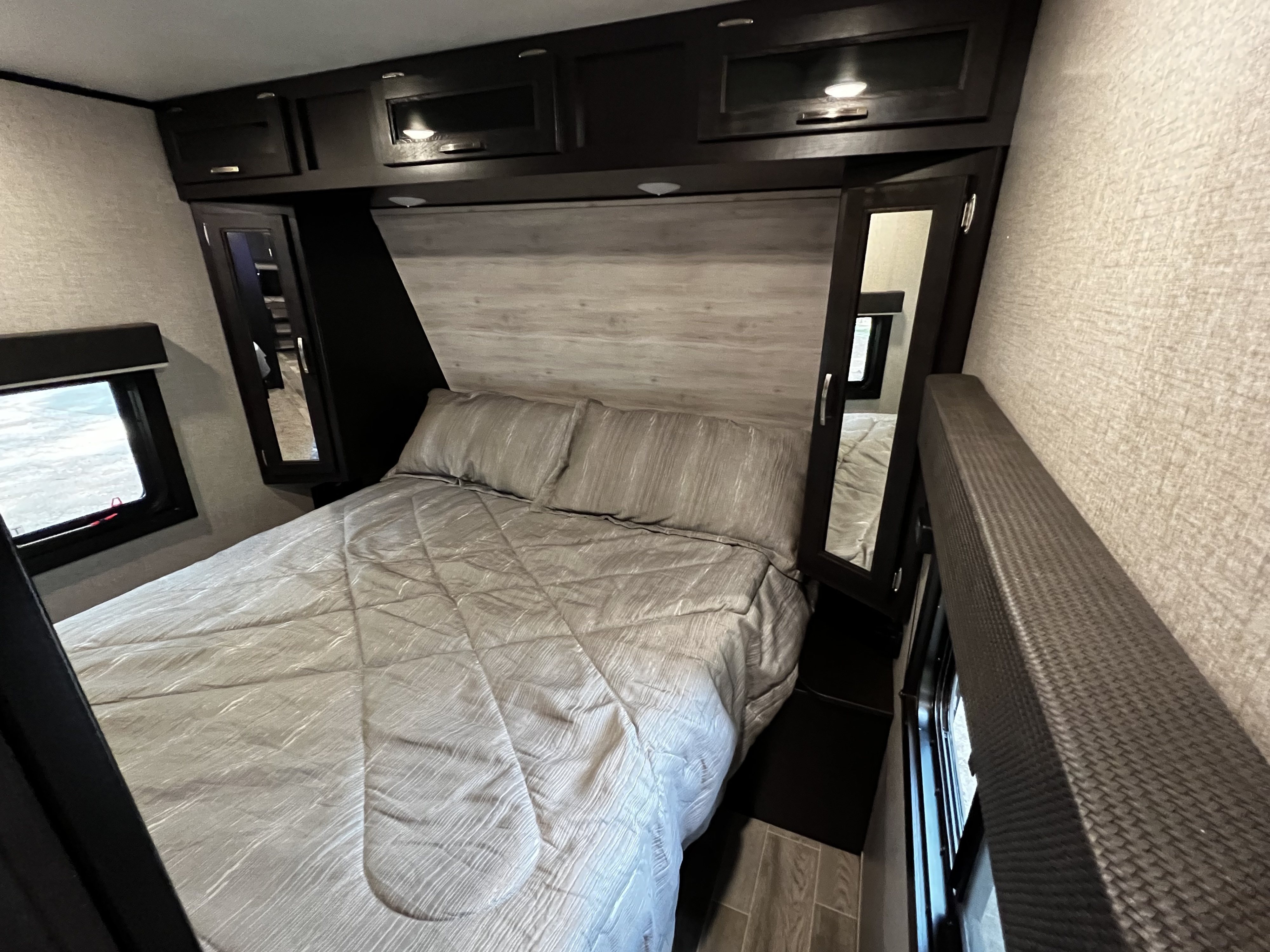 Modern RV rental bedroom: features a comfy bed with light gray linens, overhead cabinets, and compact storage. Ready for travel!. Jayco Jay Flight 2021