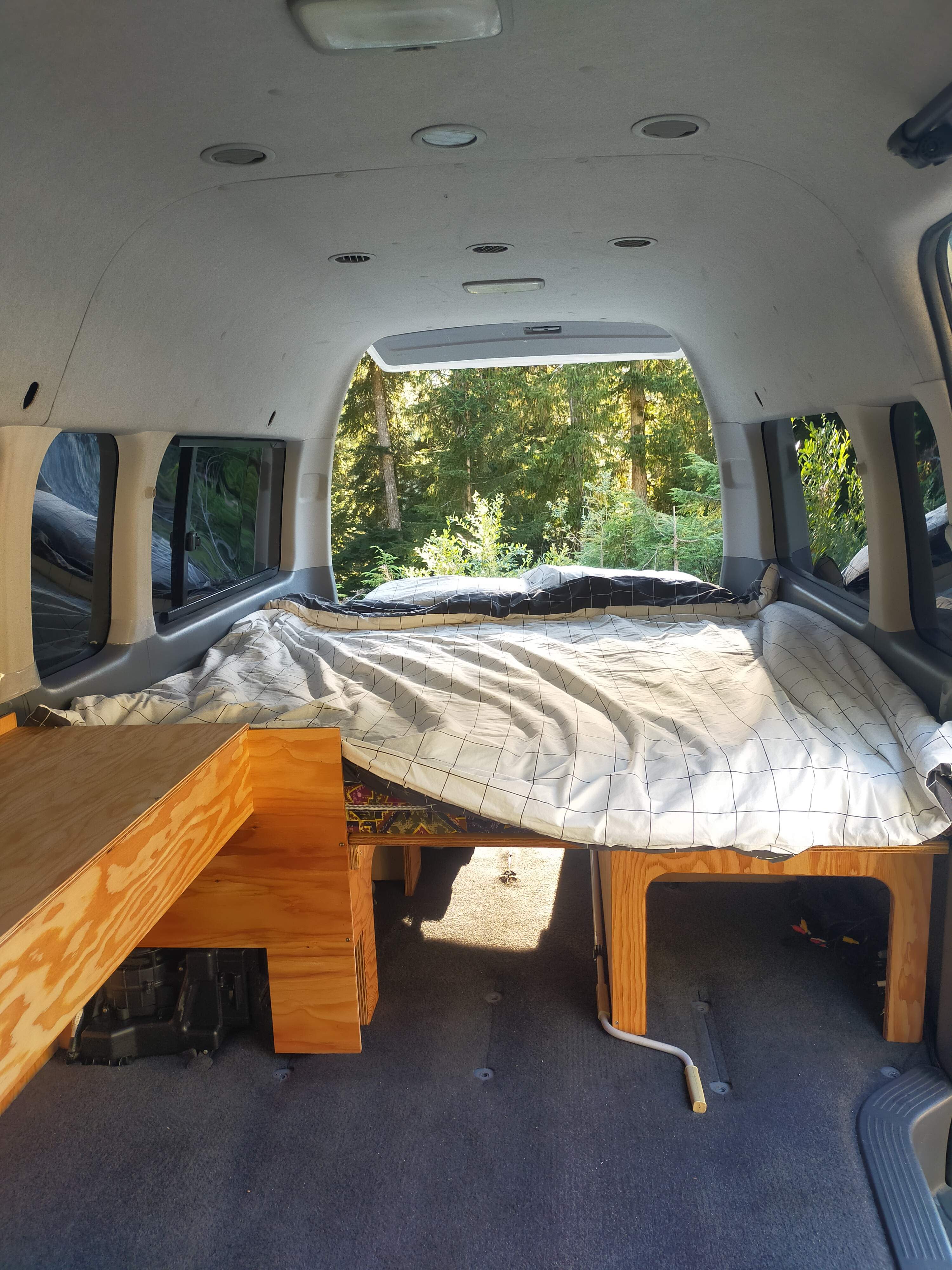RV rental interior with a cozy bed setup, offering a view of the forest.. Toyota Hiace 2005