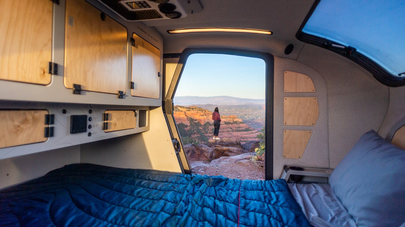 Wake up to adventure with an RV rental! Inside a cozy camper, a bed looks out to a woman on a cliff, red rock landscape.. Escapod Escapod Trailer 2023