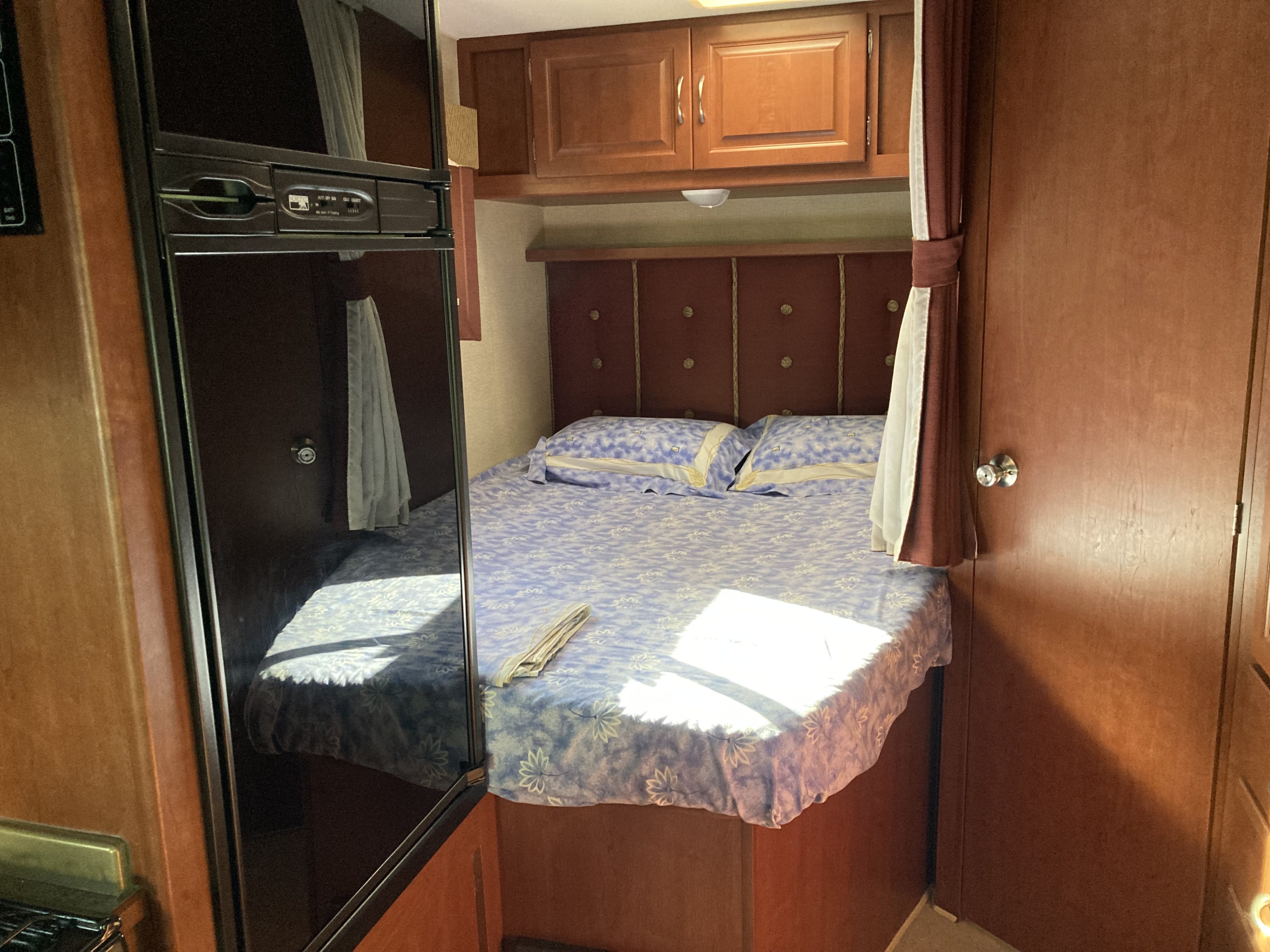Inside an RV rental, a compact bedroom features a bed with blue floral linens, wood headboard & cabinets, and a fridge.. Winnebago Minnie Winnie 2015