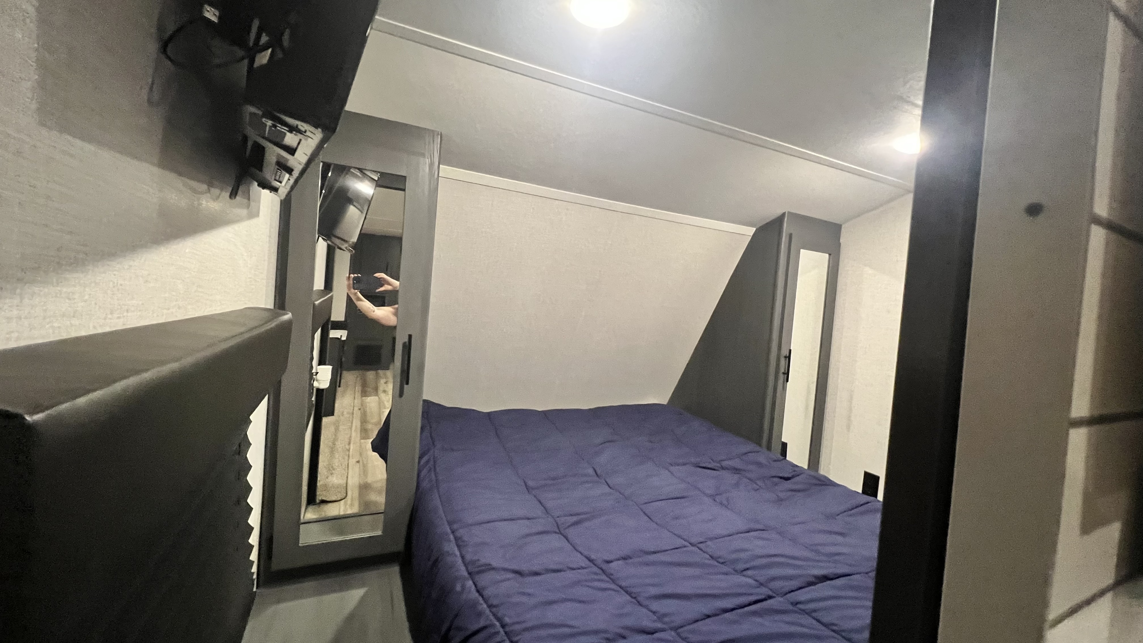 Inside an RV rental: a cozy bedroom with a blue bed, TV, and mirrored closet. A person reflects in the mirror.. Dutchmen Colorado 29BHC 2021