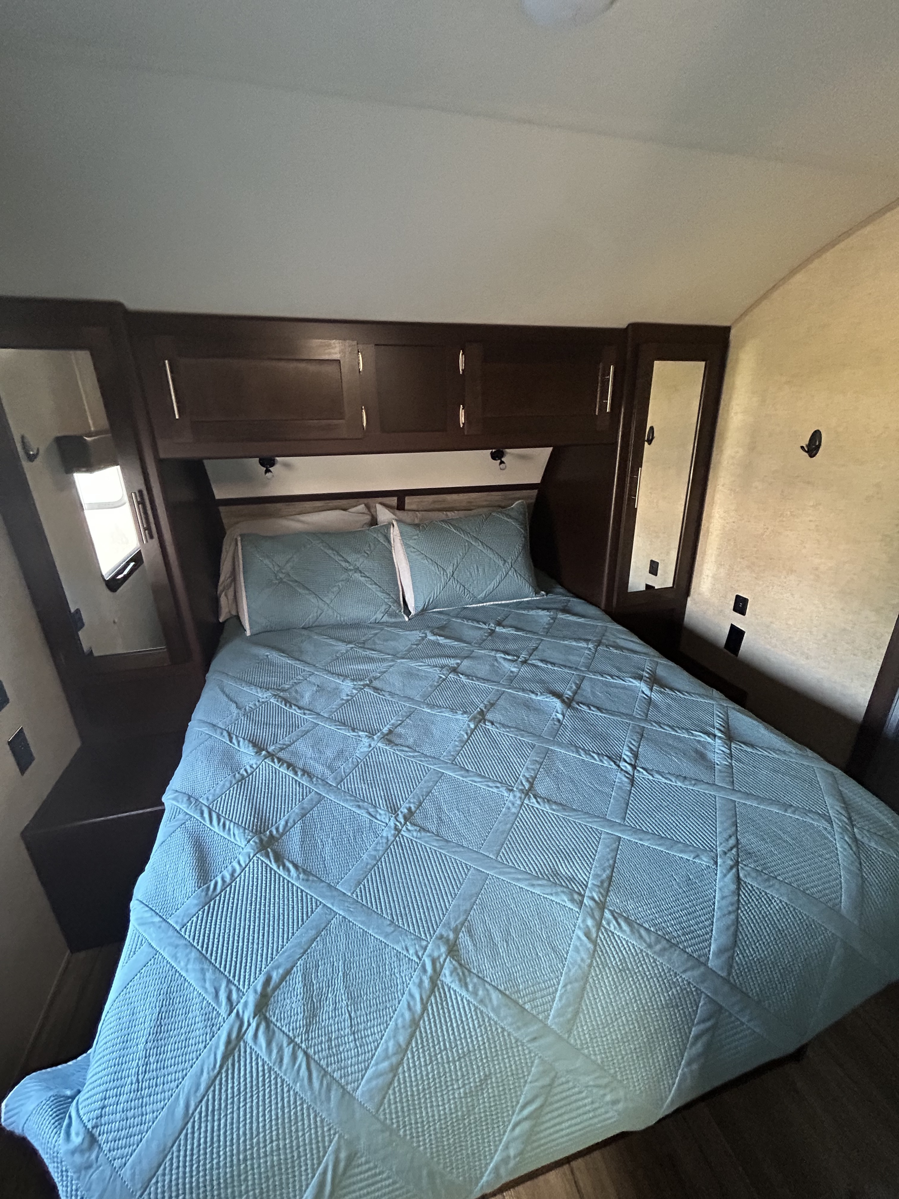 Compact RV rental bedroom featuring a blue quilted bed, overhead cabinets, and built-in mirrors.. Forest River Cherokee 2018