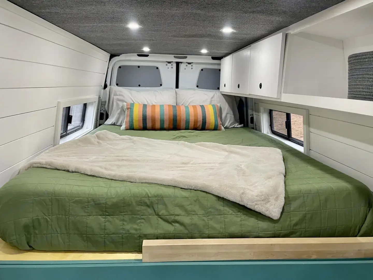 Inside this RV rental, a comfy bed with green bedding & plush blanket, white shiplap walls, and practical overhead storage.. Mercedes Sprinter 2020