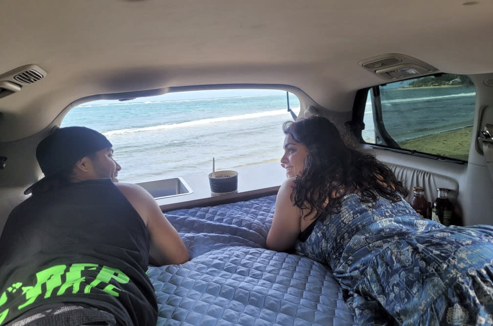 A couple smiles, lying inside their RV rental with a fantastic ocean and beach view from the open rear of the camper van.. Toyota Sienna 2008