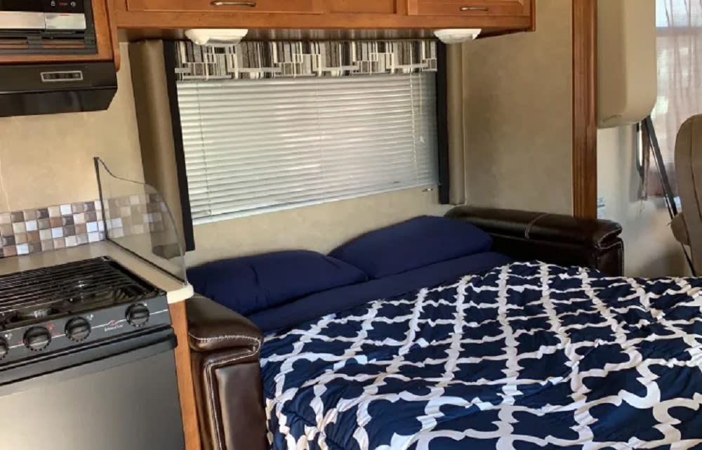 RV rental interior showing a comfortable pull-out bed, window with blinds, and compact kitchen area.. Coachmen Pursuit 2014