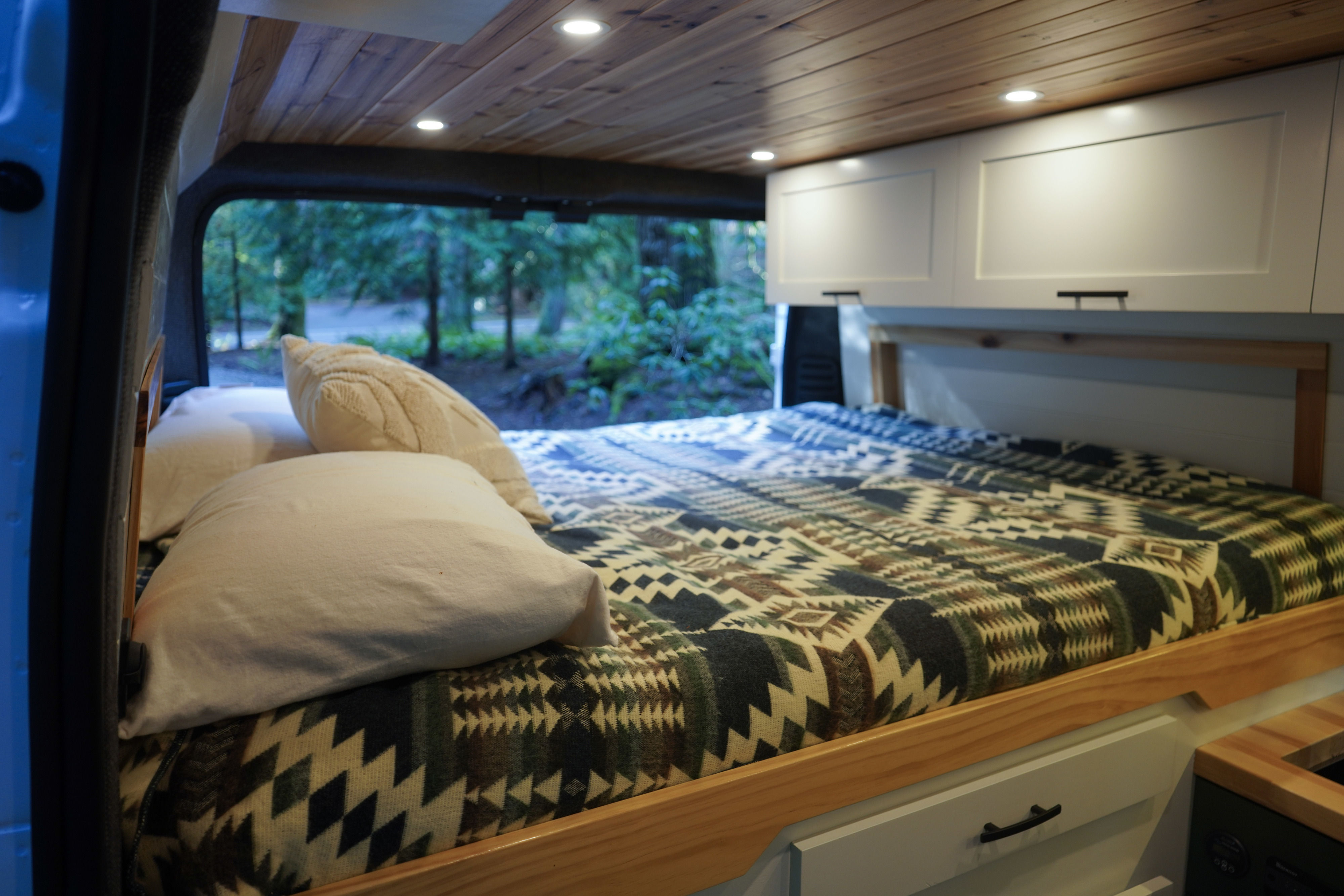 Cozy RV rental interior with a bed, wooden ceiling, cabinets, and a scenic forest view through the back window.. Dodge RAM Promaster 1500 2020