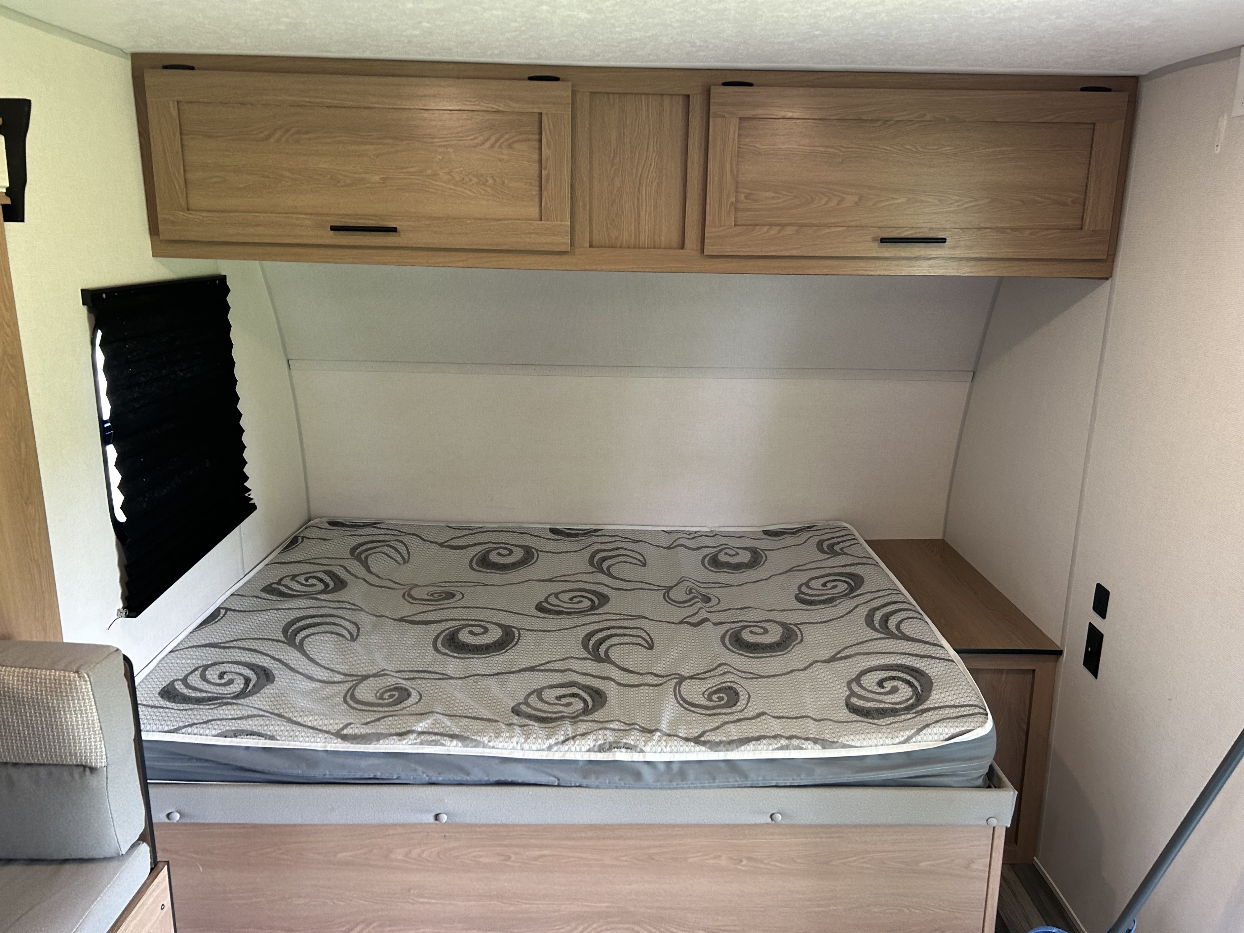 RV rental bedroom interior showing a bed with patterned mattress, wooden overhead cabinets, and a window with black blinds.. Avenger LT 2022