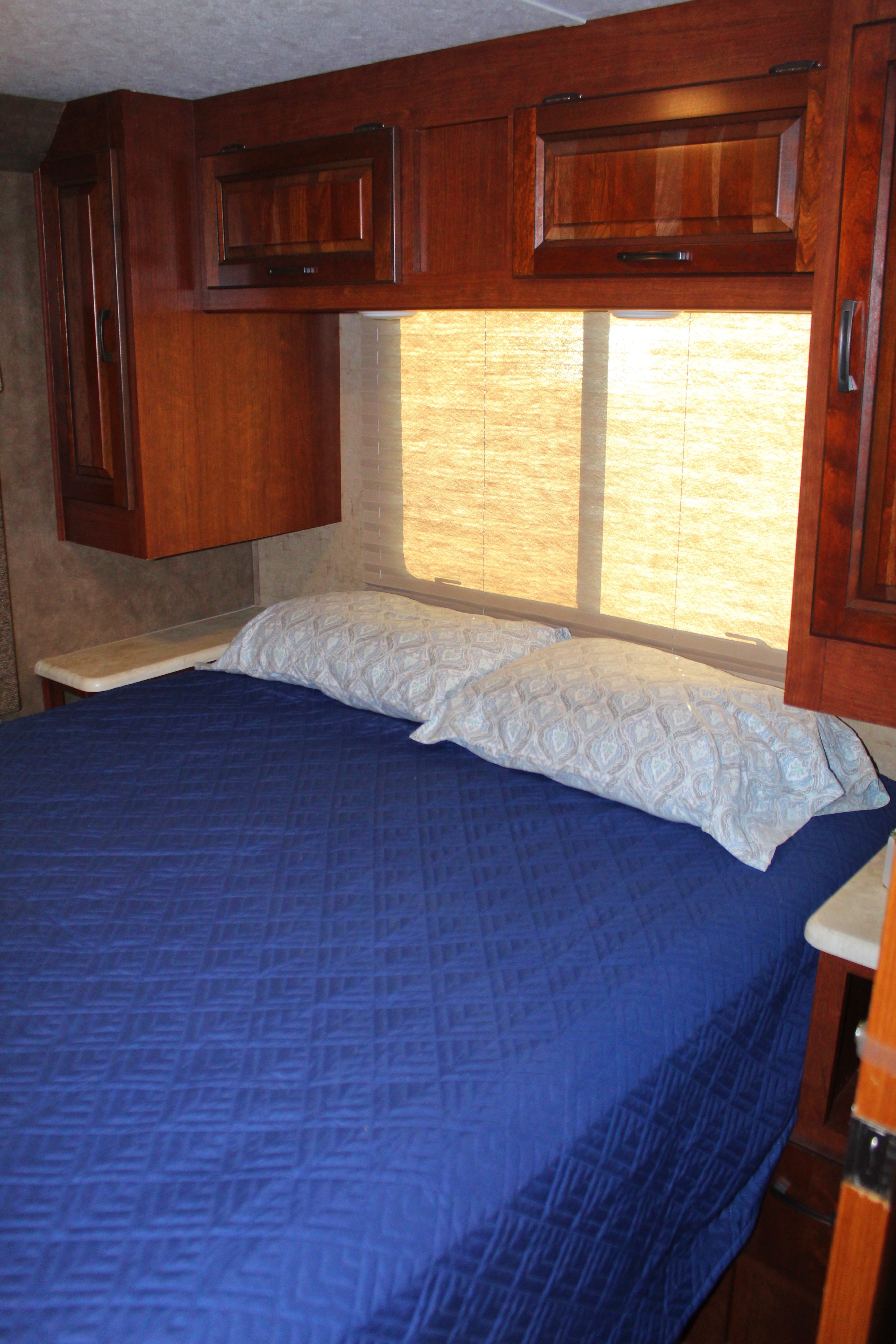 Inside this RV rental, a comfy bedroom setup with a blue quilted bed, patterned pillows, and dark wood overhead cabinets by a bright window.. Forest River Sunseeker 2015