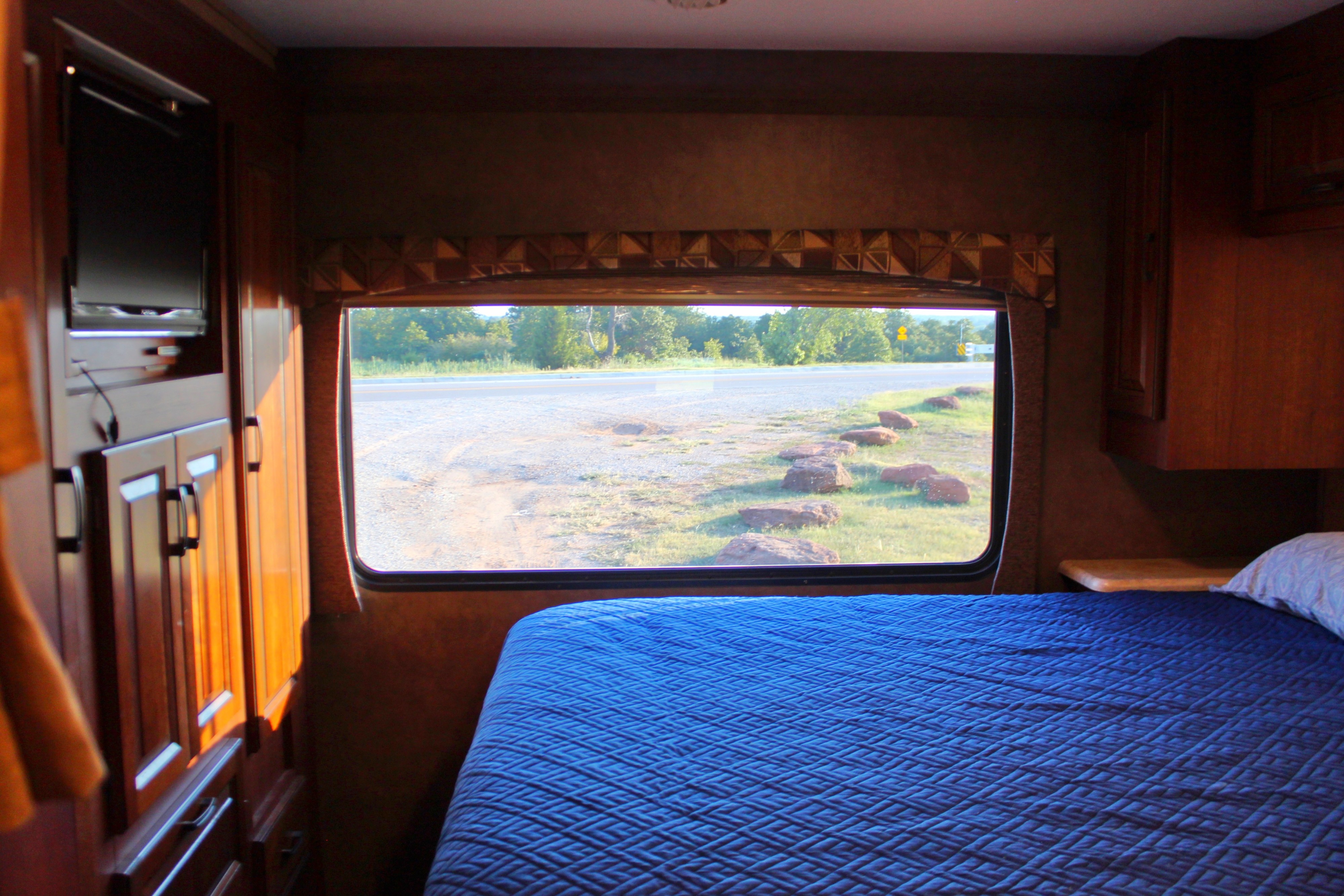Cozy RV bedroom with a blue quilted bed and a large window view of a quiet road. Perfect for your RV rental journey.. Forest River Sunseeker 2015