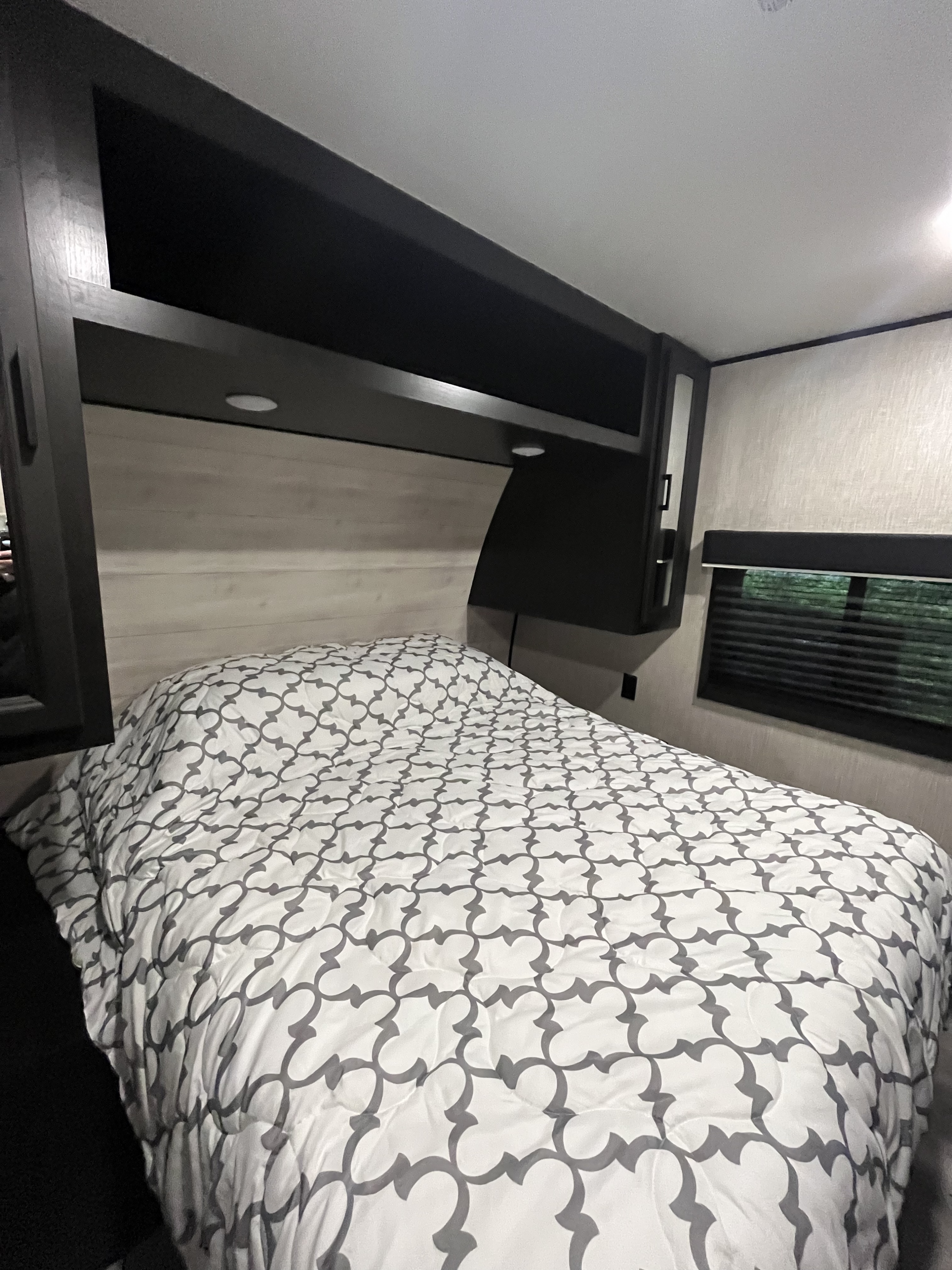 Cozy RV rental bedroom featuring a white & gray patterned comforter, dark built-in overhead storage, and a window with blinds.. Jayco Jay Flight 2023
