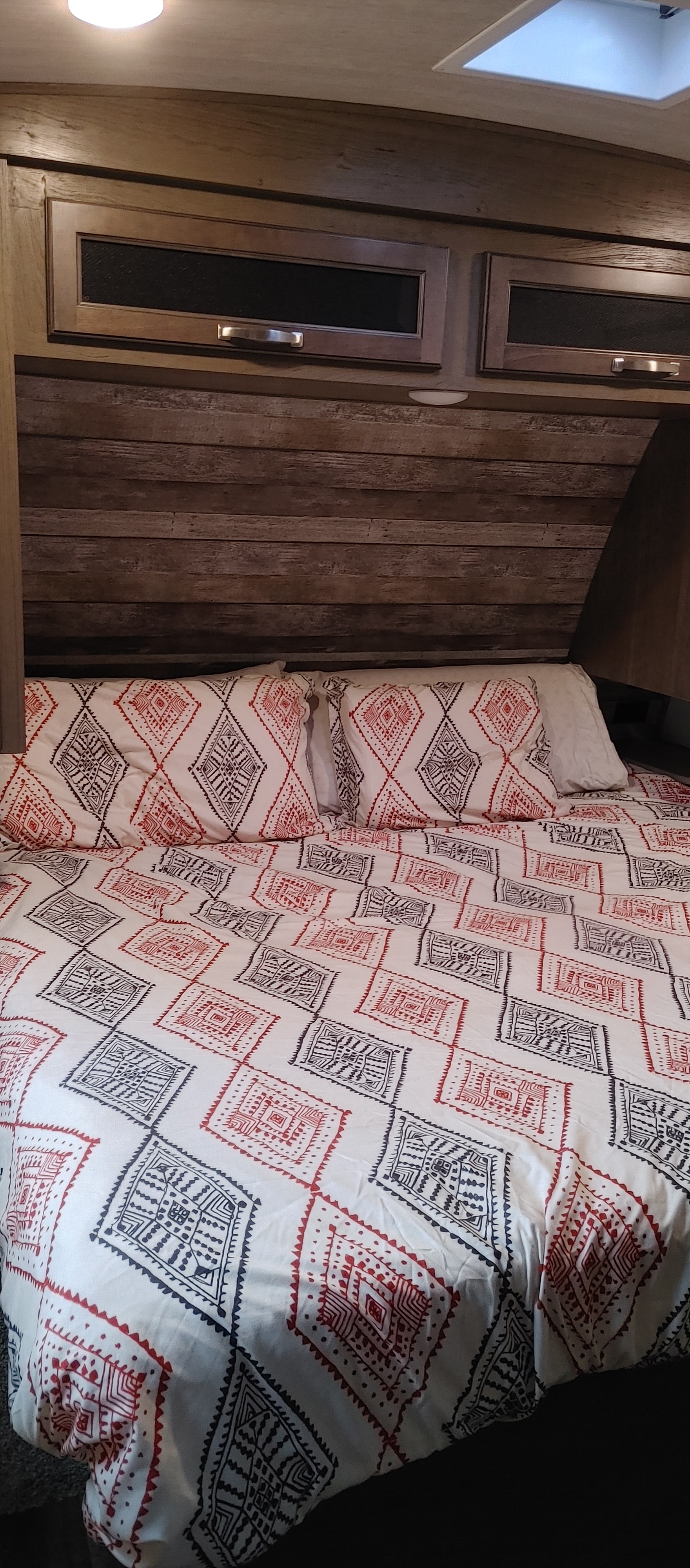 RV rental bedroom: a bed with red and dark geometric bedding, wooden headboard, and overhead cabinets.. Keystone RV Cougar 2021