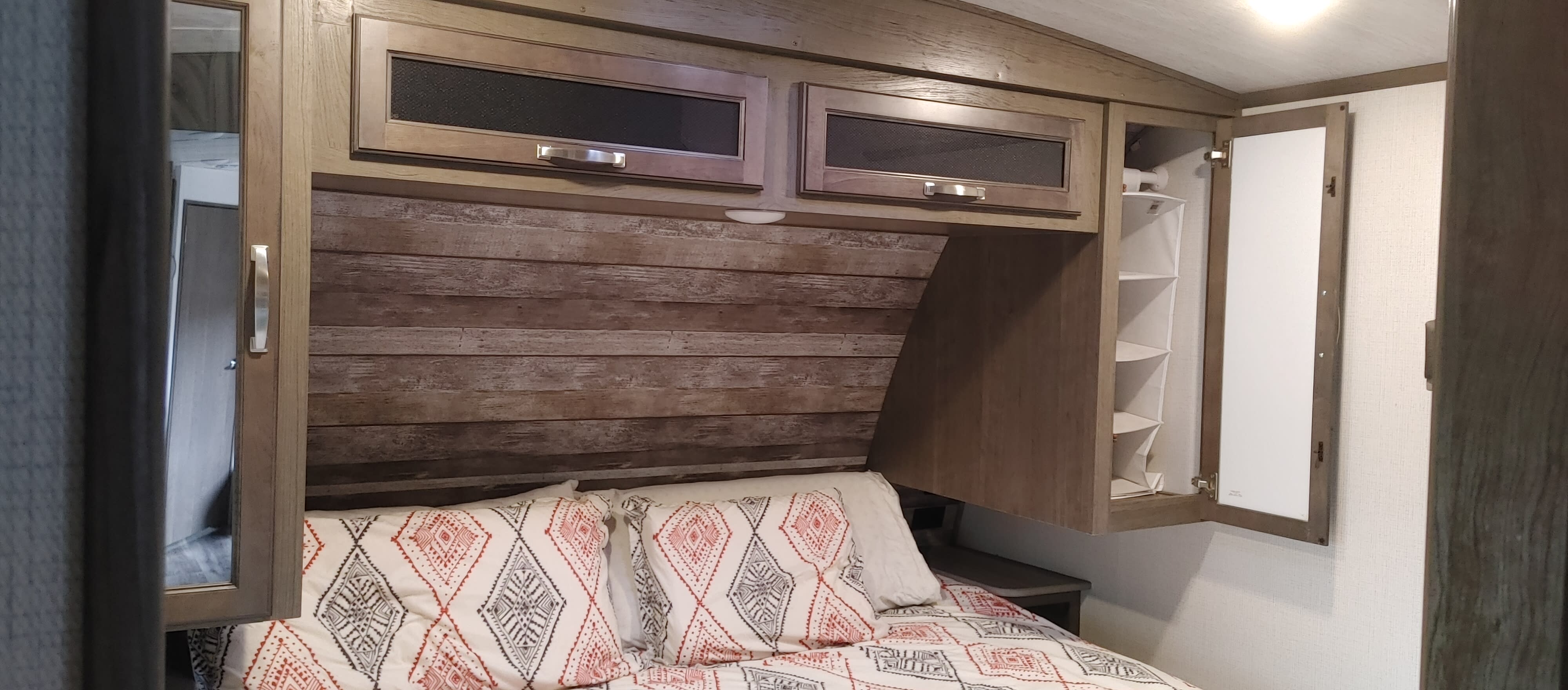 RV rental bedroom featuring a cozy bed, wood-paneled wall, and overhead storage, including an open closet with shelves.. Keystone RV Cougar 2021