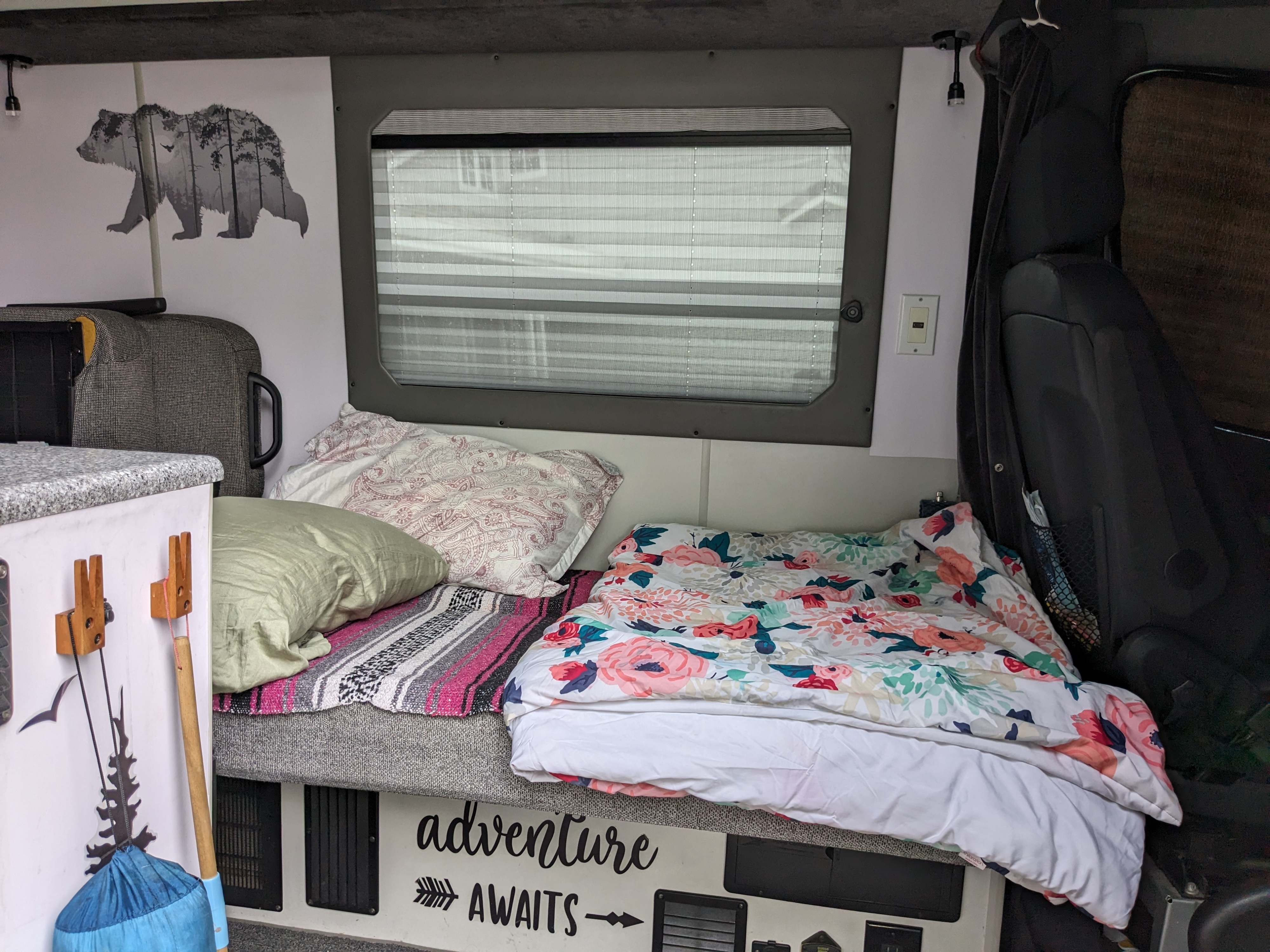 Inviting RV rental interior featuring a floral-patterned bed, bear wall art, and an "adventure AWAITS" sign.. Sportsmobile Long Body 2013