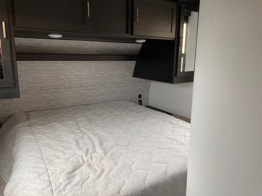 Inside this RV rental, a compact bedroom features a grey bed, dark overhead cabinets, and geometric patterned walls.. Keystone RV Outback Ultra-Lite 2022