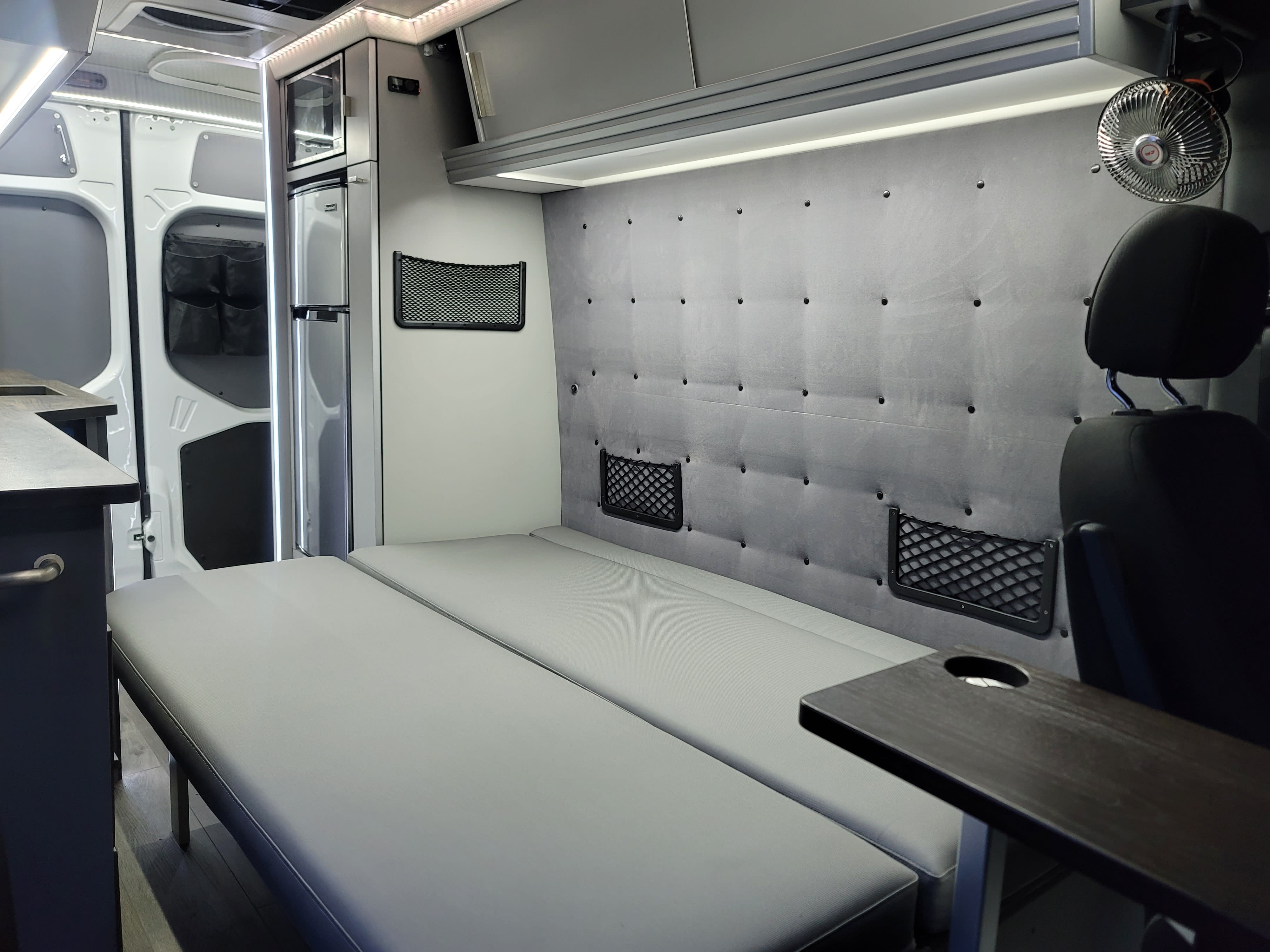 Interior of a grey rv rental camper van with a convertible bed, built-in storage nets, compact fridge, and fan.. Mercedes 2500 2022