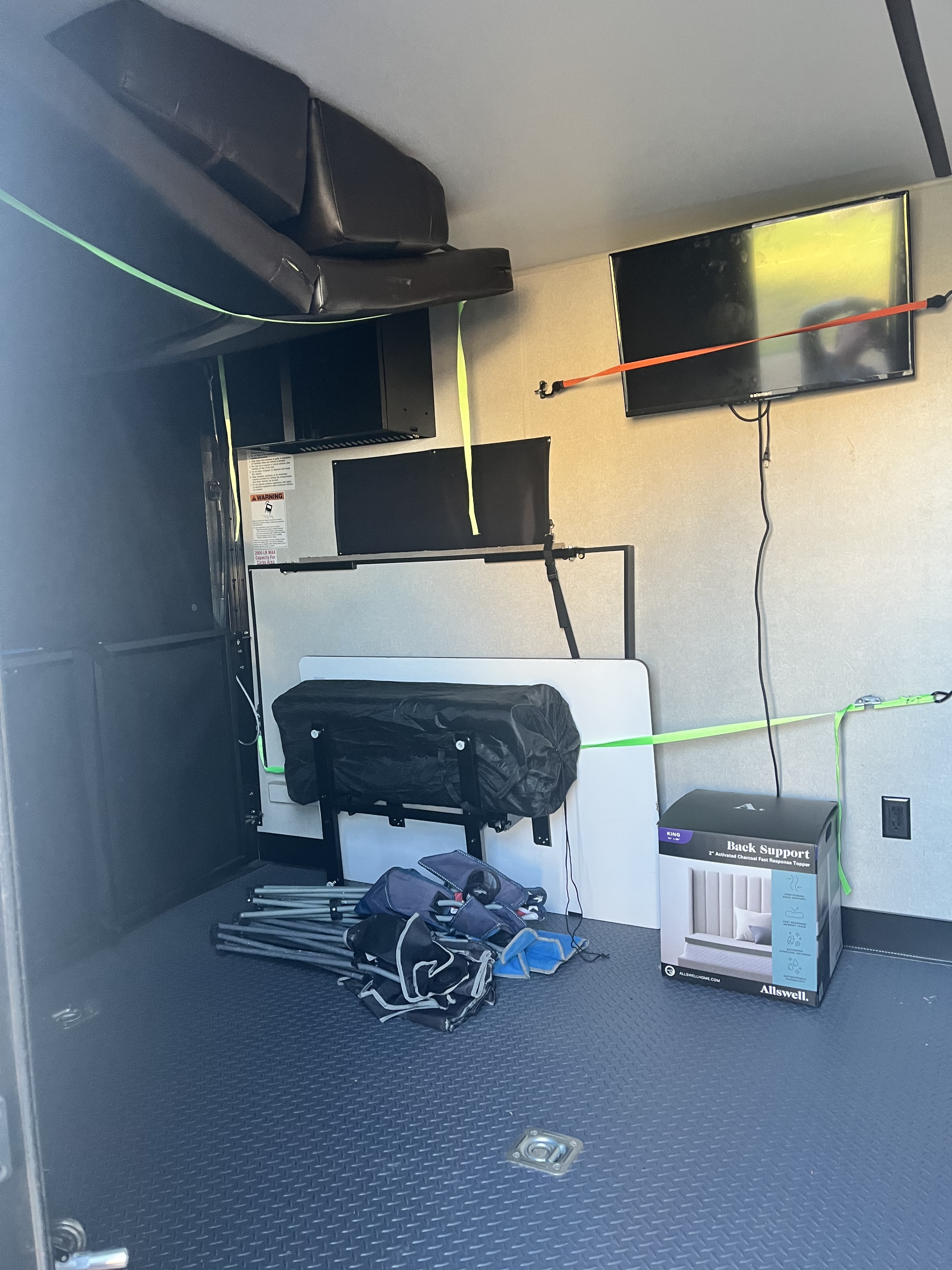 Inside an RV rental's organized cargo area, showing overhead folded beds, mounted TVs, and secured tables/chairs on a textured floor.. Forest River XLR Micro Boost Toy Hauler 2020
