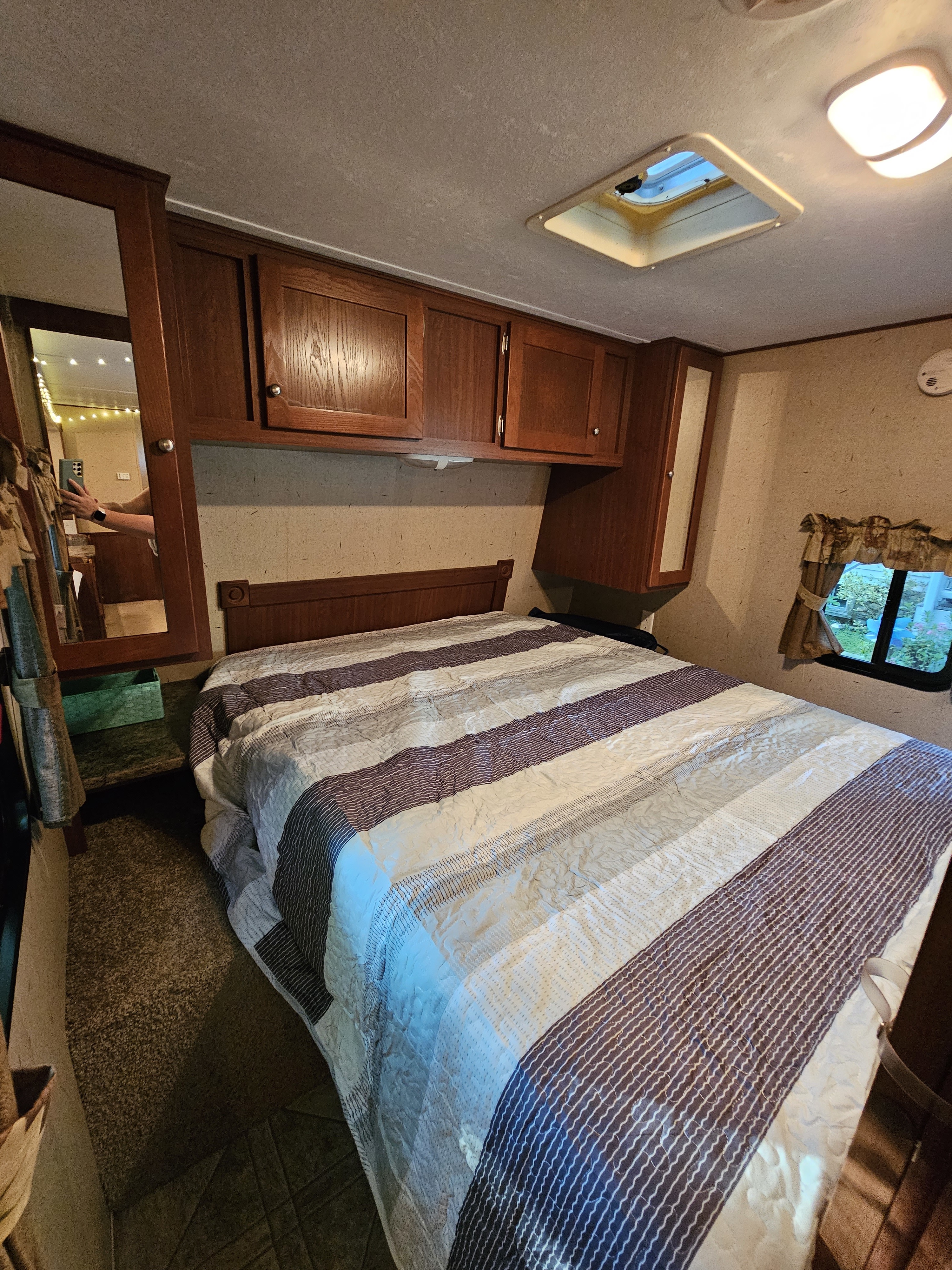 Cozy RV rental bedroom featuring striped bedding, overhead storage cabinets, a mirror, and a window with outdoor view.. Crossroads RV Zinger 2010