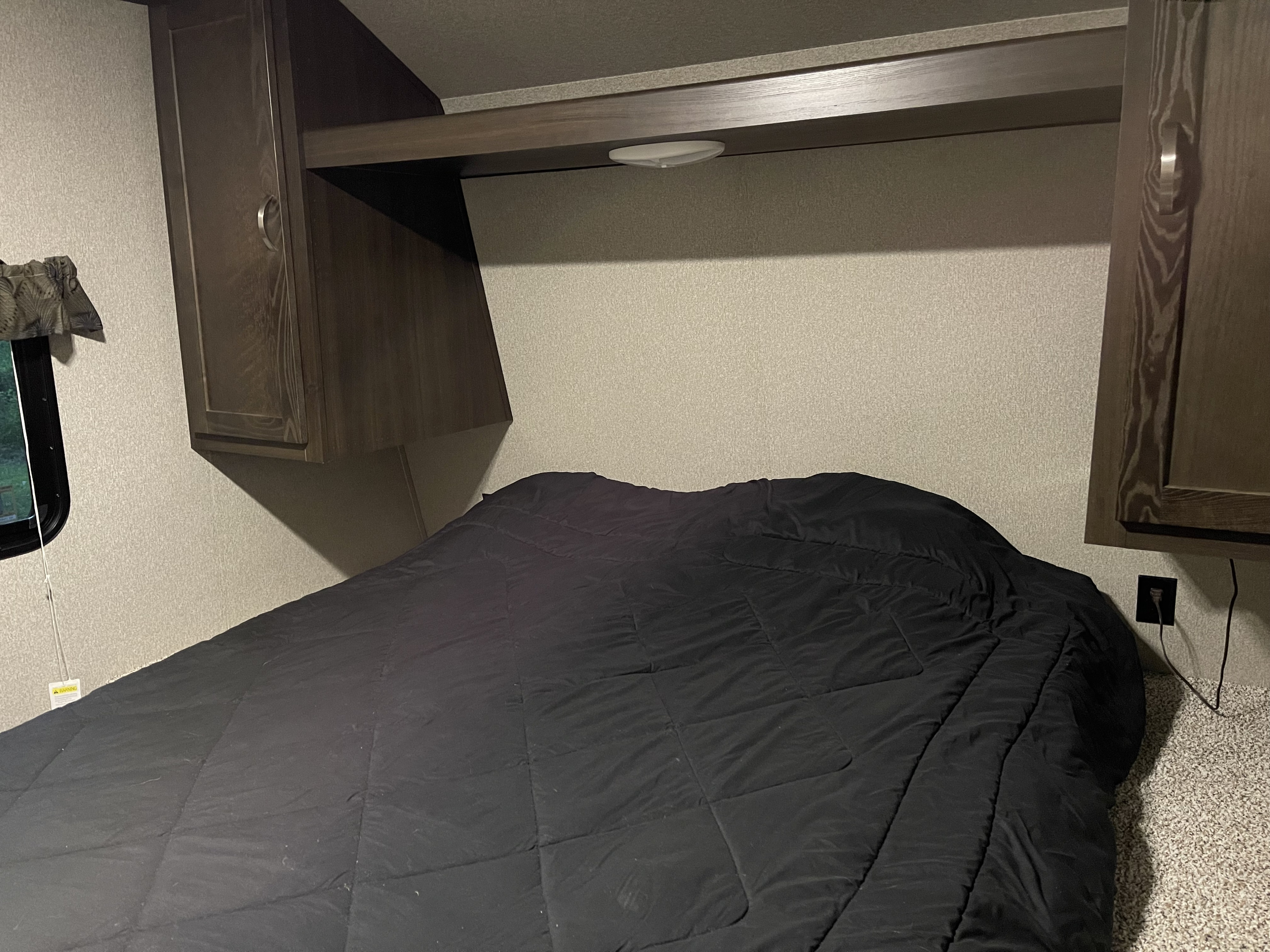 Bedroom interior of an RV rental, showing a bed with a dark comforter, overhead cabinets, and a window.. Keystone RV Springdale 2018