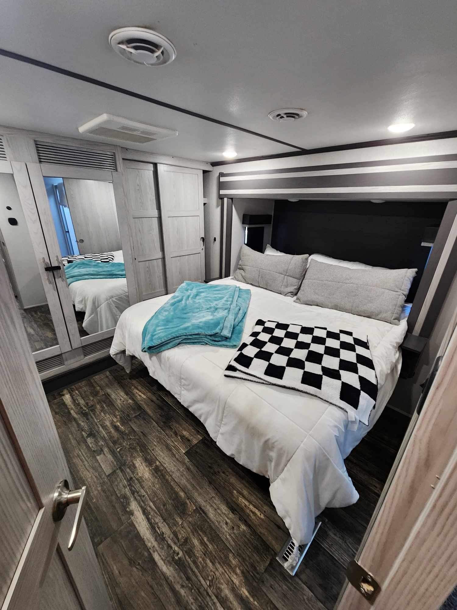 This RV rental bedroom features a comfortable bed with blue and checkered blankets, a mirrored closet, and dark wood floors.. Keystone RV Fuzion Toy Hauler 2021