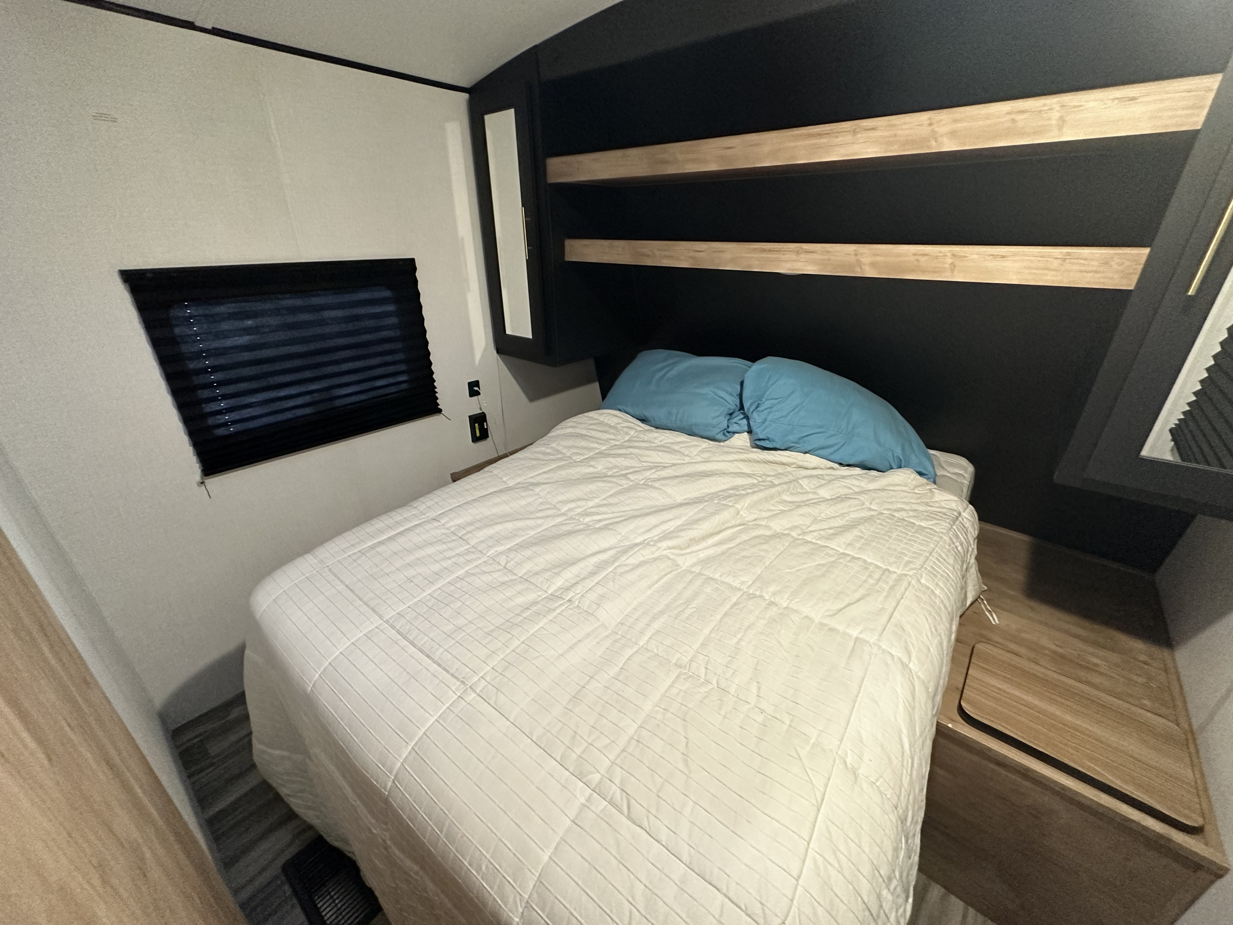Inside a cozy RV rental bedroom: bed with blue pillows, overhead shelves, window, and storage. Ideal for travel.. Keystone RV Springdale 2023