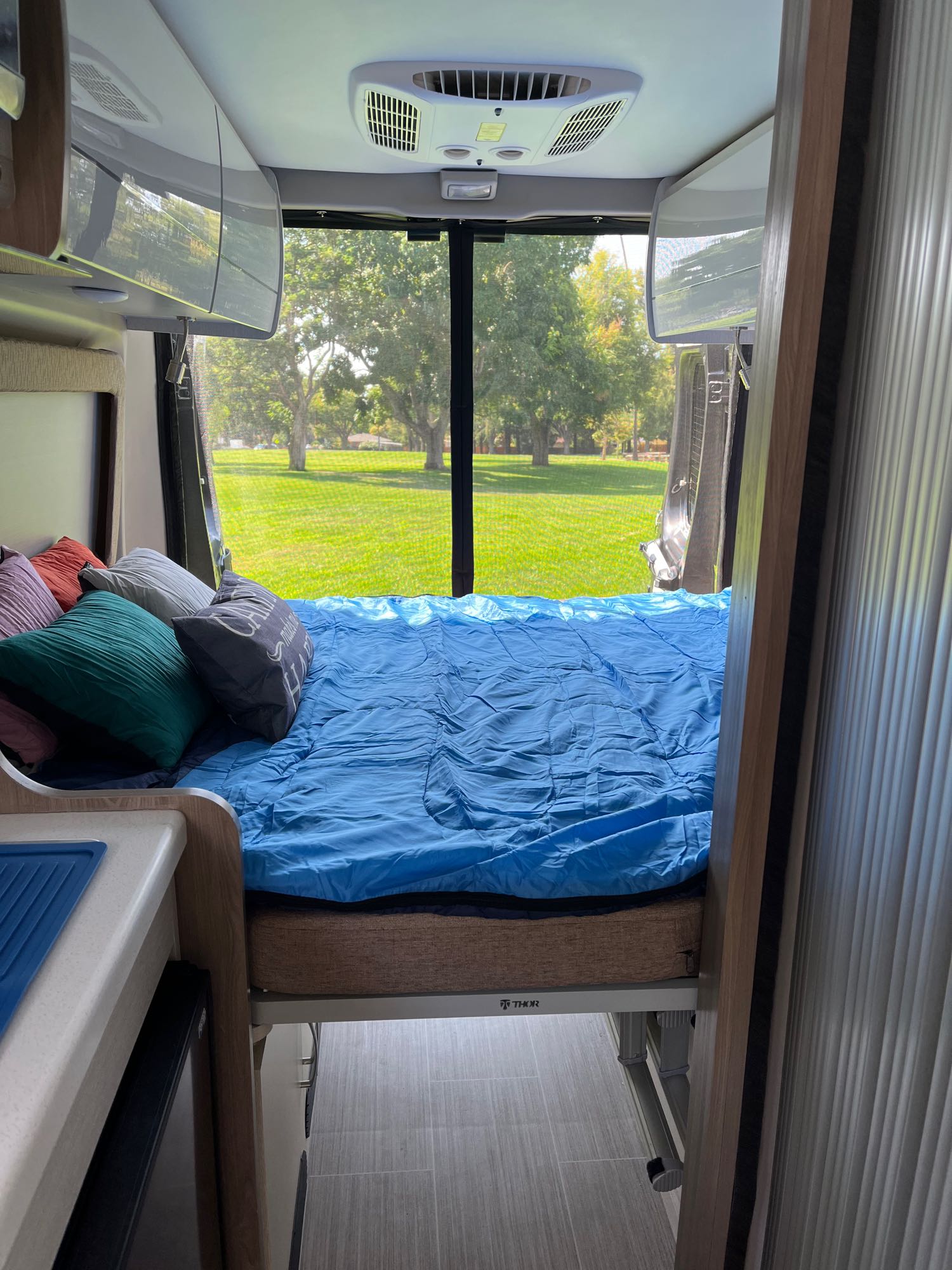 Inside an rv rental: a neatly made bed with a blue comforter offers a serene park view. Perfect for cozy camping and travel adventures.. Thor Tellaro 2022